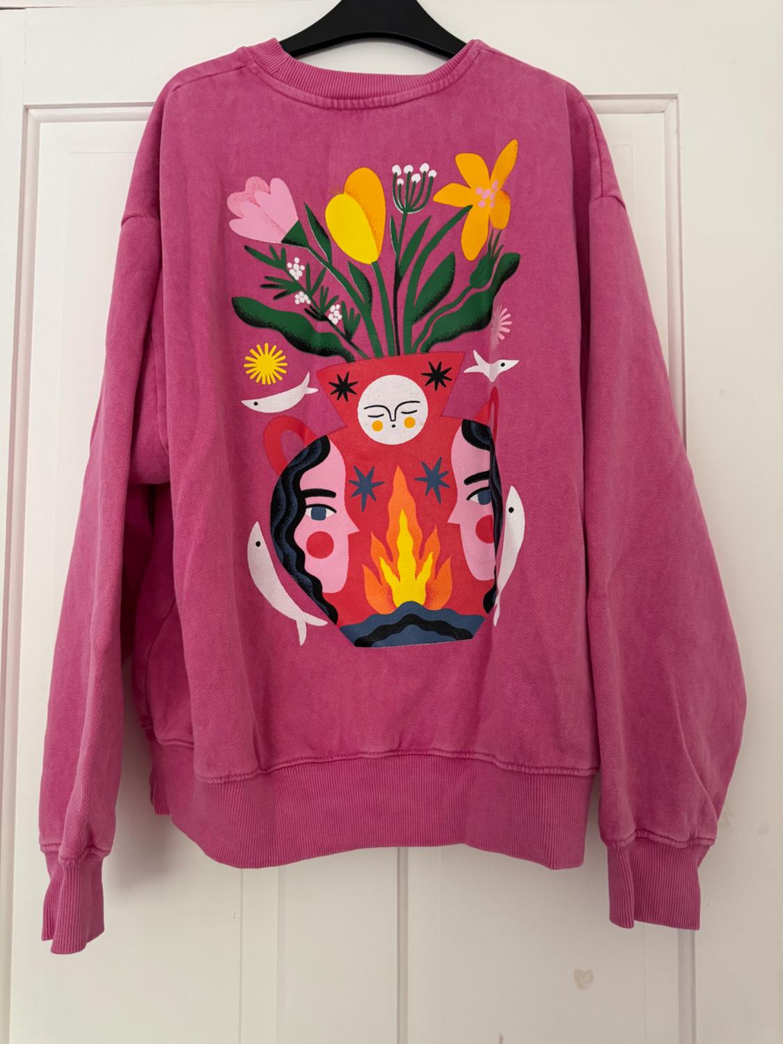 Pink sweatshirt hanging on a white door. It features a creative and colourful print of a red vase with faces, fire, flowers, and white fish.
