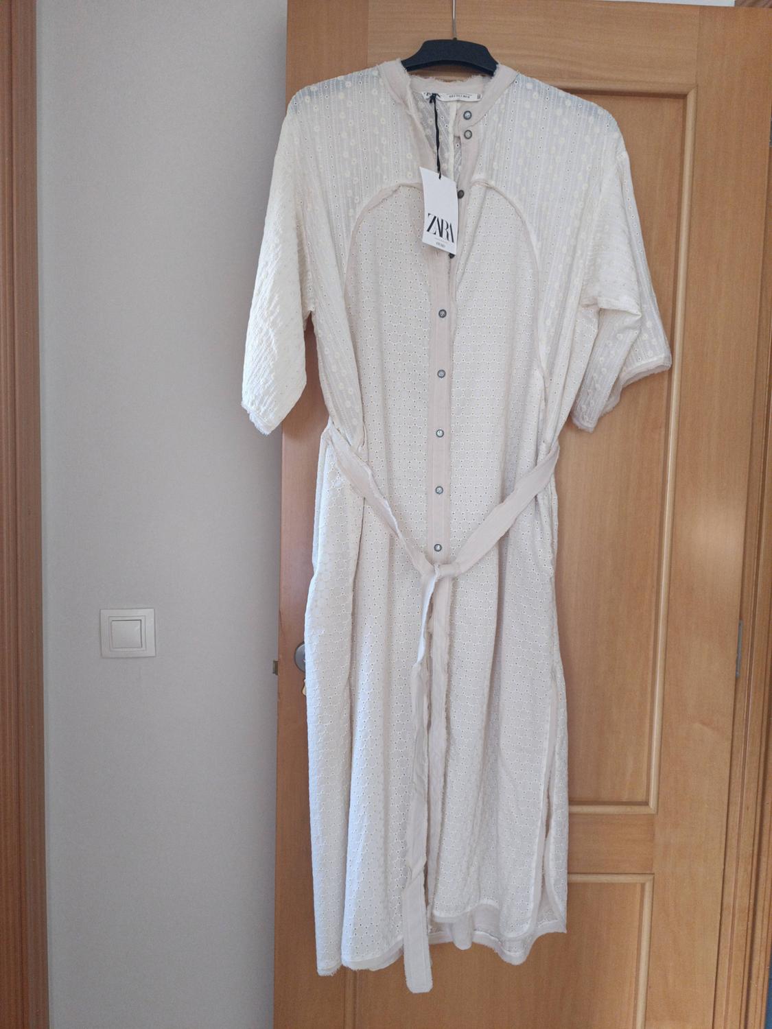 White short-sleeved button-through long dress with a belt, hanging on a wooden door. Label with the text ZARA.