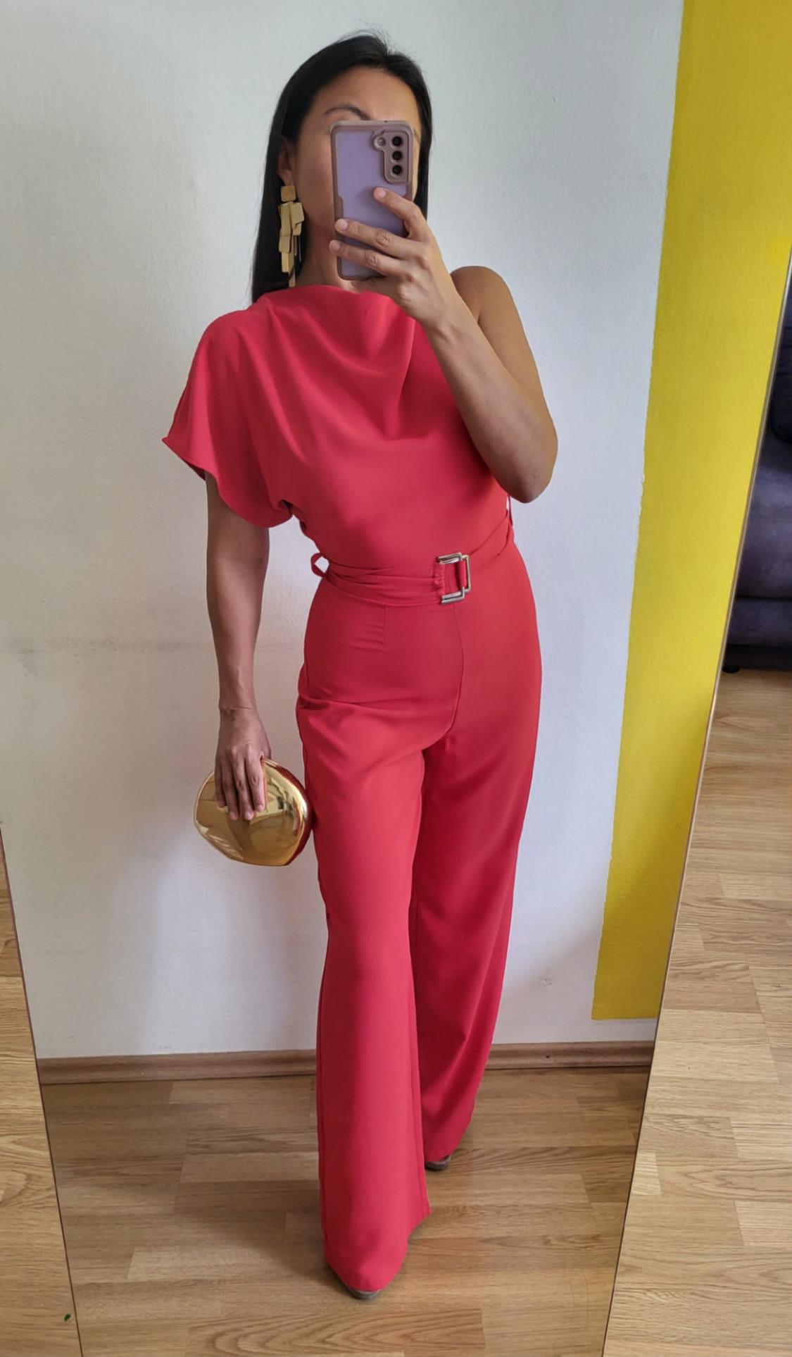 Woman wearing a red, one-shoulder jumpsuit, with a belt and gold earrings, holding a gold handbag, in front of a mirror.