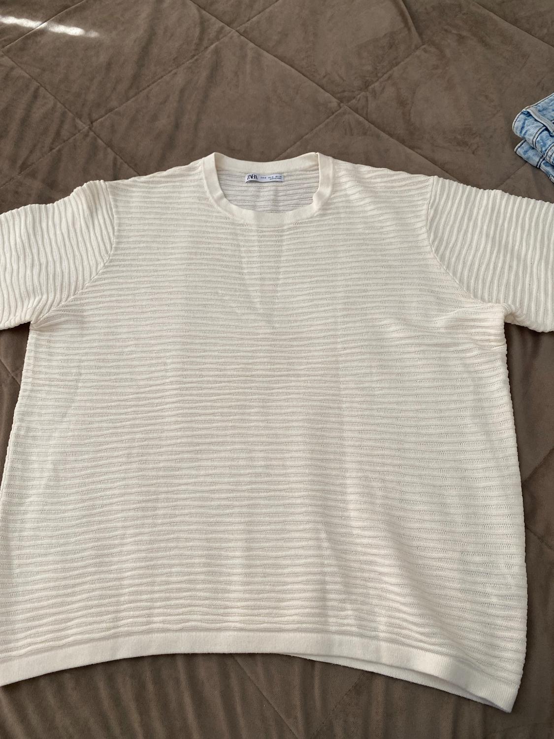 White short-sleeved T-shirt with a striped texture on a brown quilt. ZARA label visible at the neck.