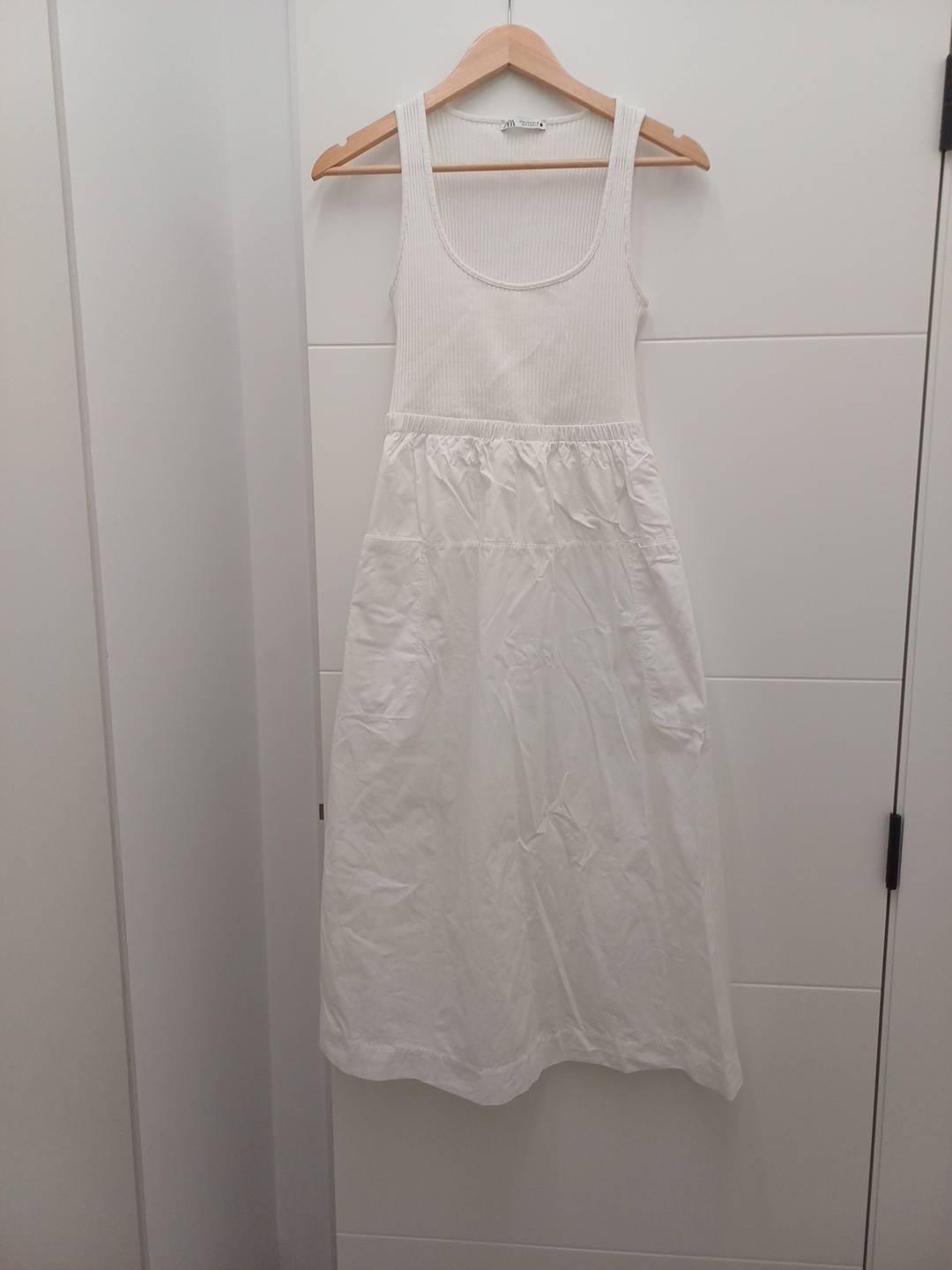 White dress hanging on a wooden hanger on a white door. Ribbed bodice with straps and skirt with pockets.