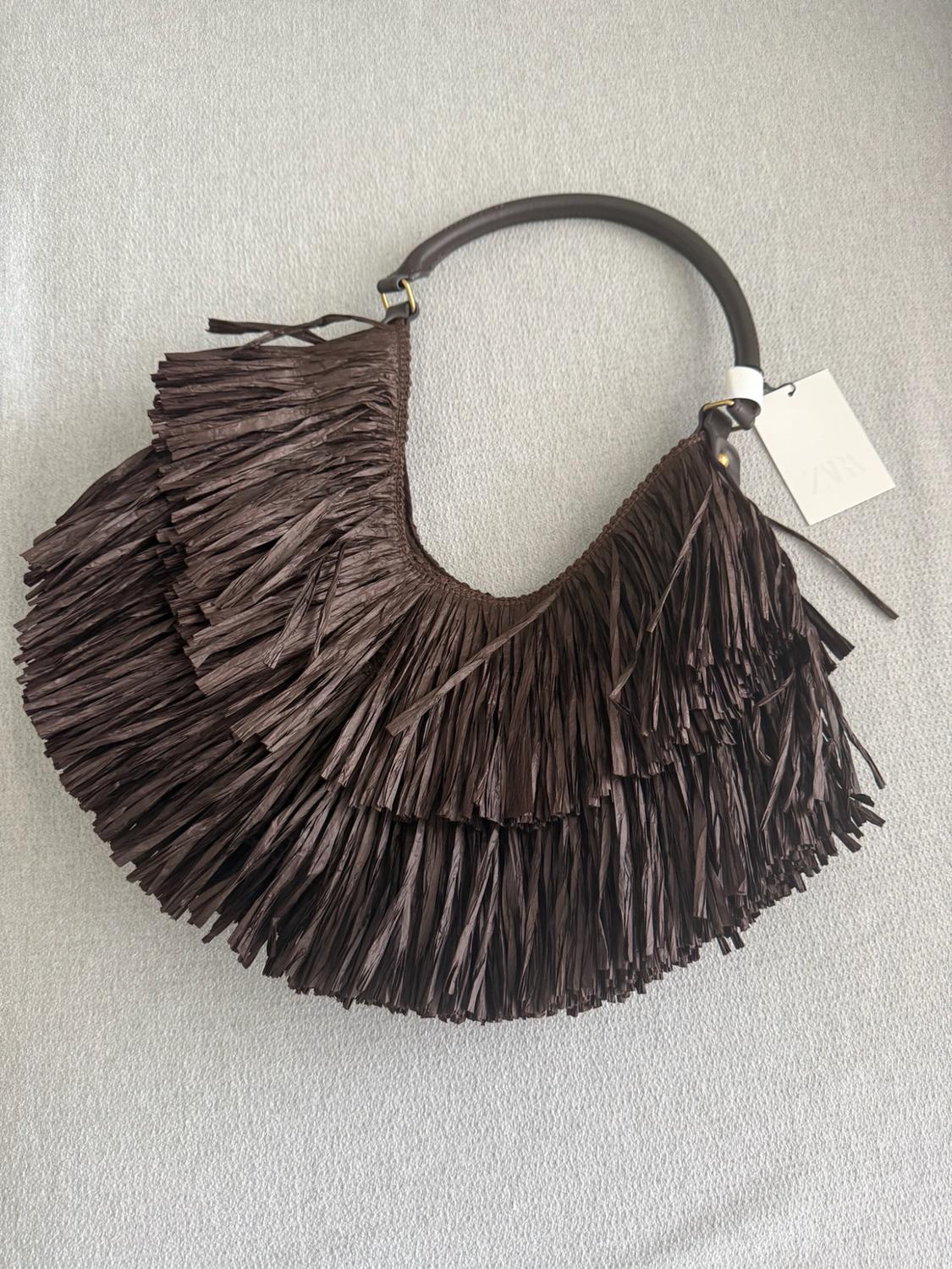 Dark brown fringed bag with a curved handle on a light grey fabric surface. White tag with the text ZARA.