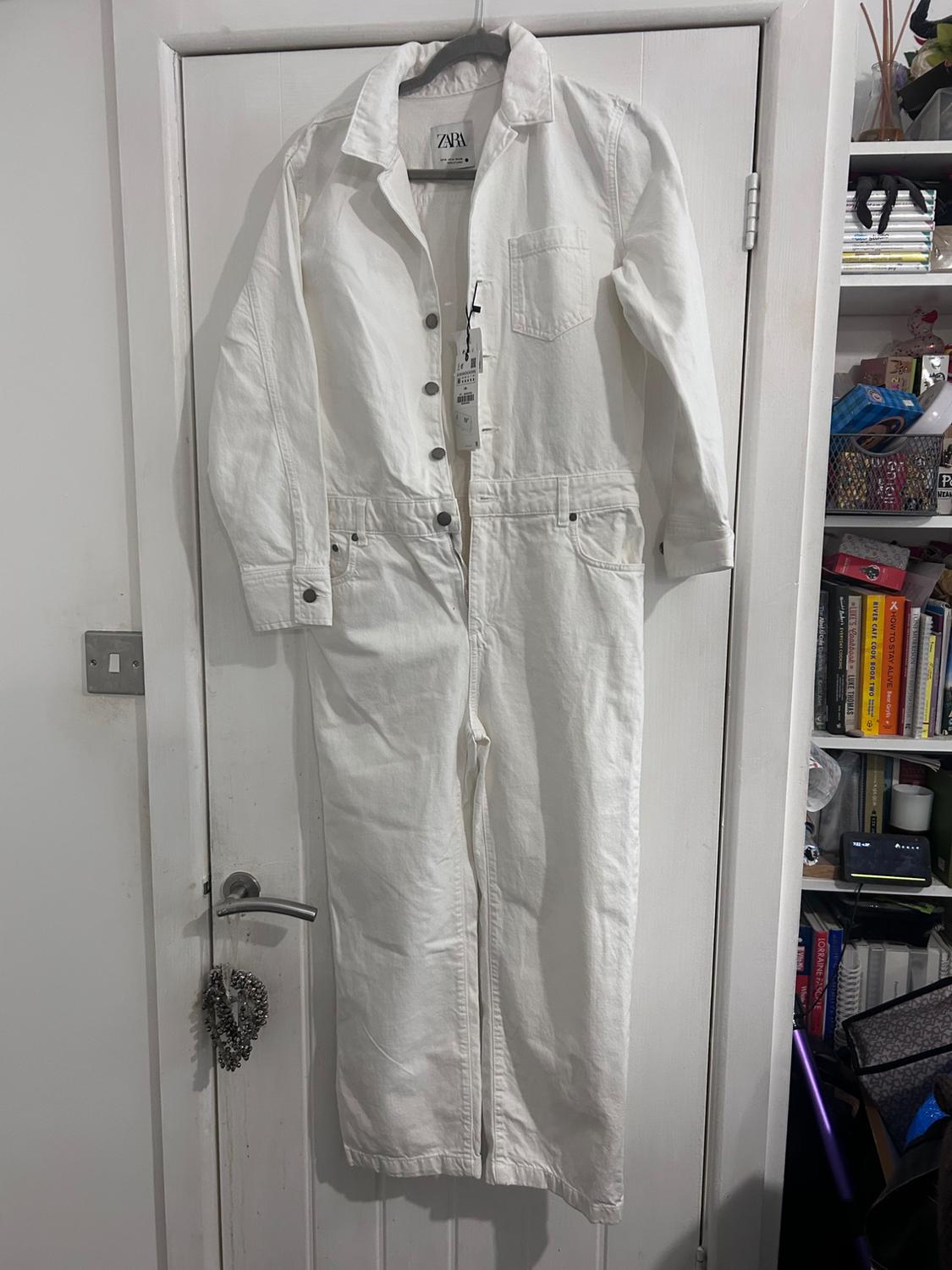 White jumpsuit hanging on a white door, with a visible Zara label. Background with a bookshelf full of books and various objects.