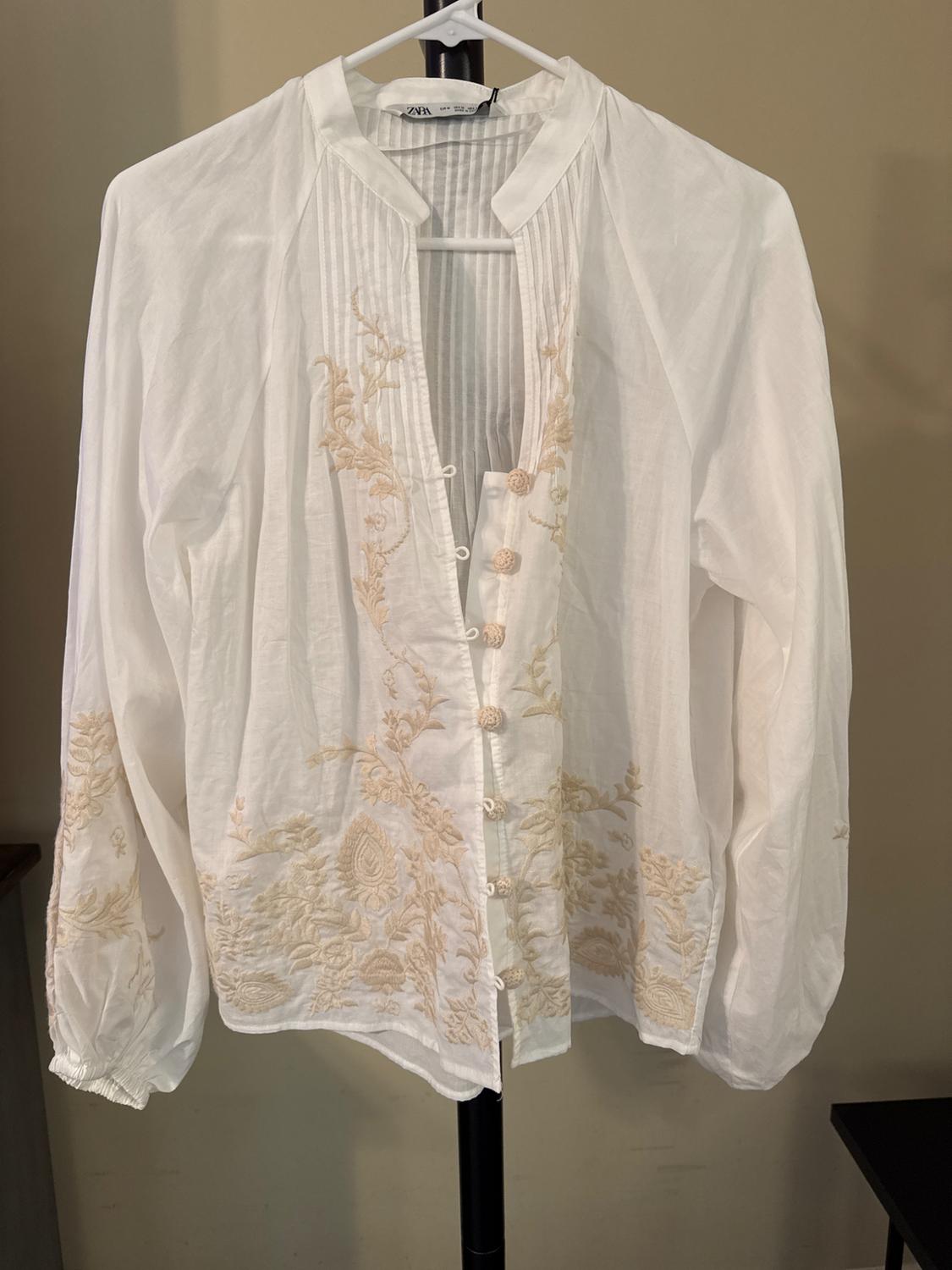 Front view of a white blouse with beige floral embroidery, round neckline, central button closure, and long sleeves.