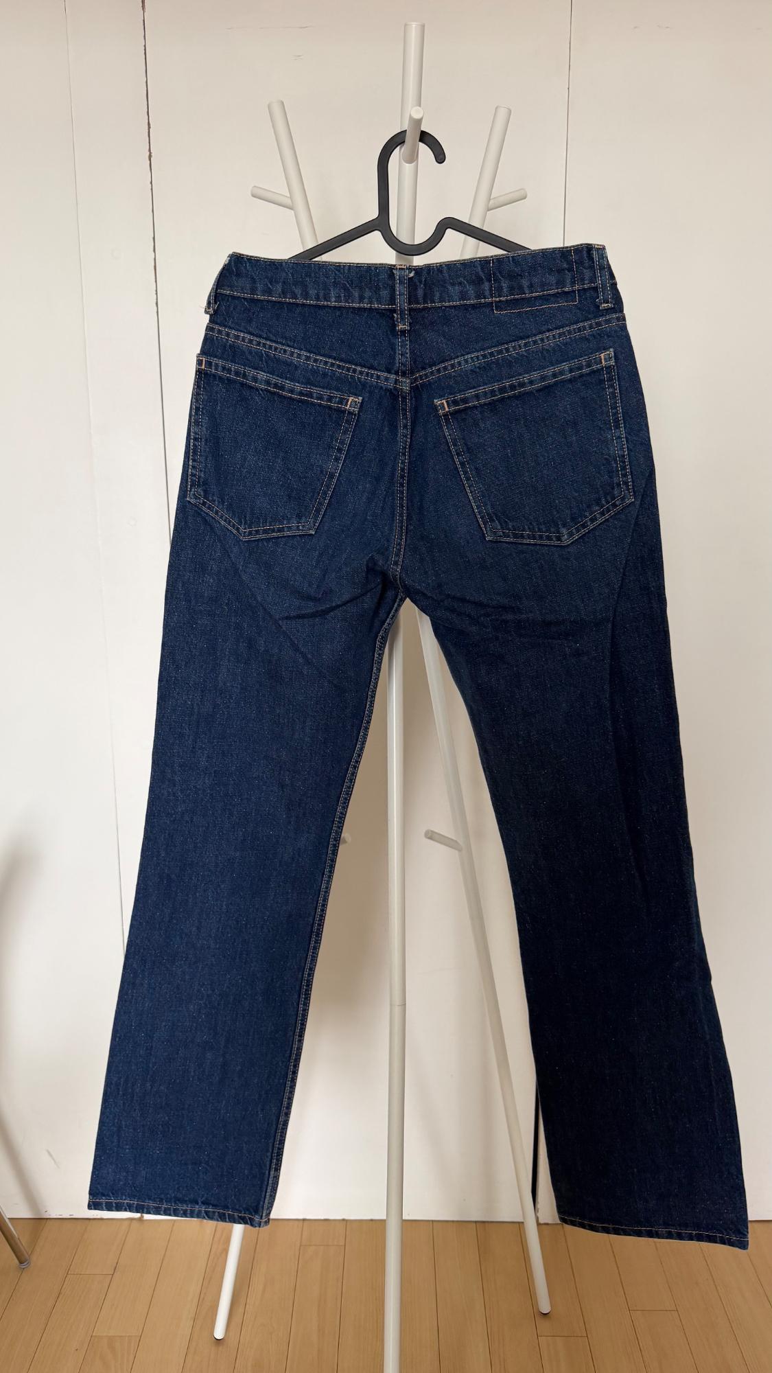 Rear view of a pair of dark blue jeans hanging on a white clothes rack. In the background, a white wall and light wood flooring.