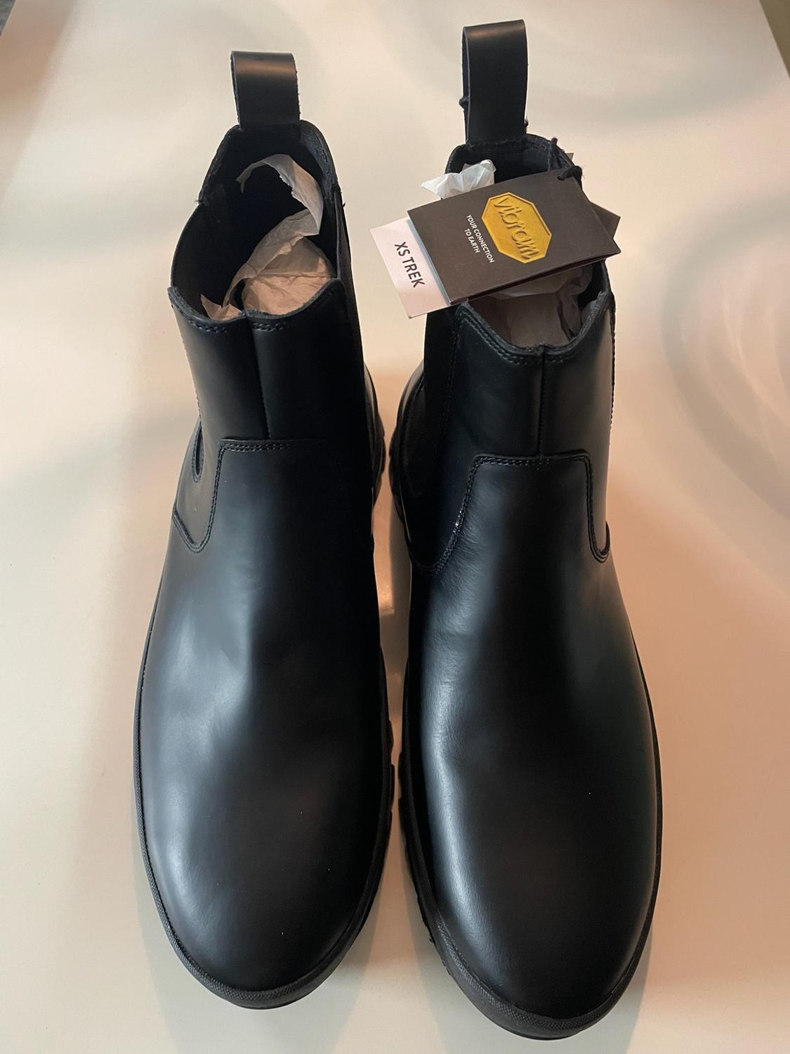 Black Chelsea-style boots on a light background. They have labels bearing the text “Vibram YOUR CONNECTION TO EARTH” and “XS TREK”.