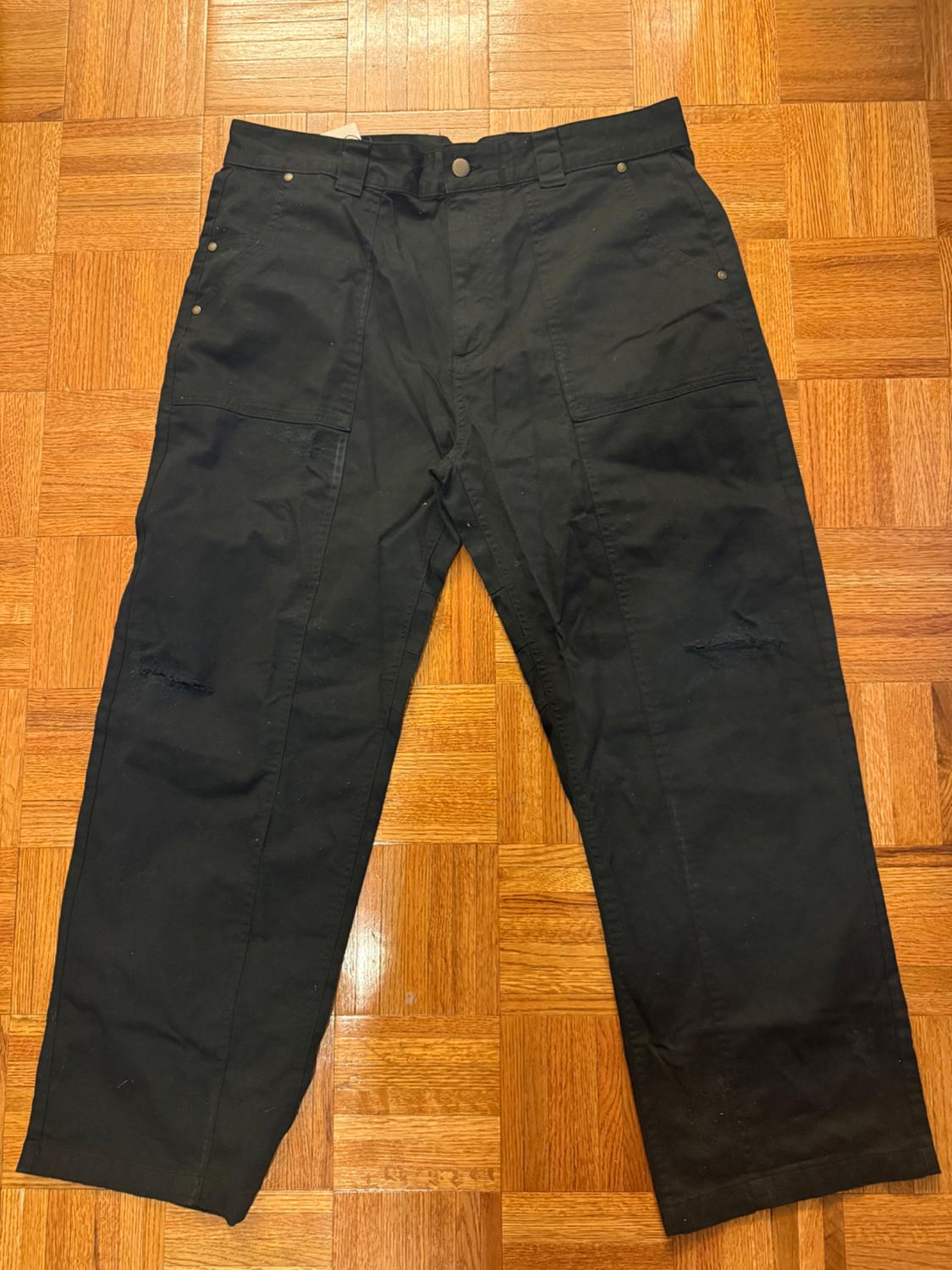 Black, straight-cut trousers with pockets and metallic buttons, laid out on a light wooden floor.