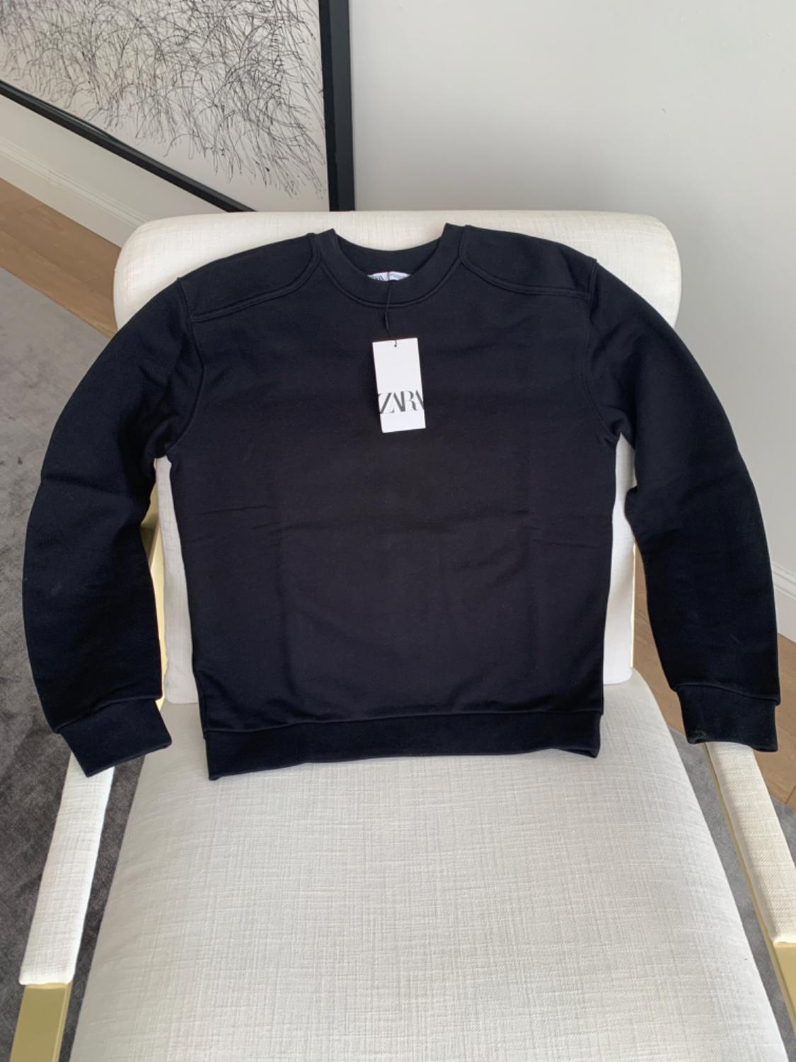 Black sweatshirt with a "ZARA" tag hanging off it, on a white chair. Background with a white wall and abstract painting.