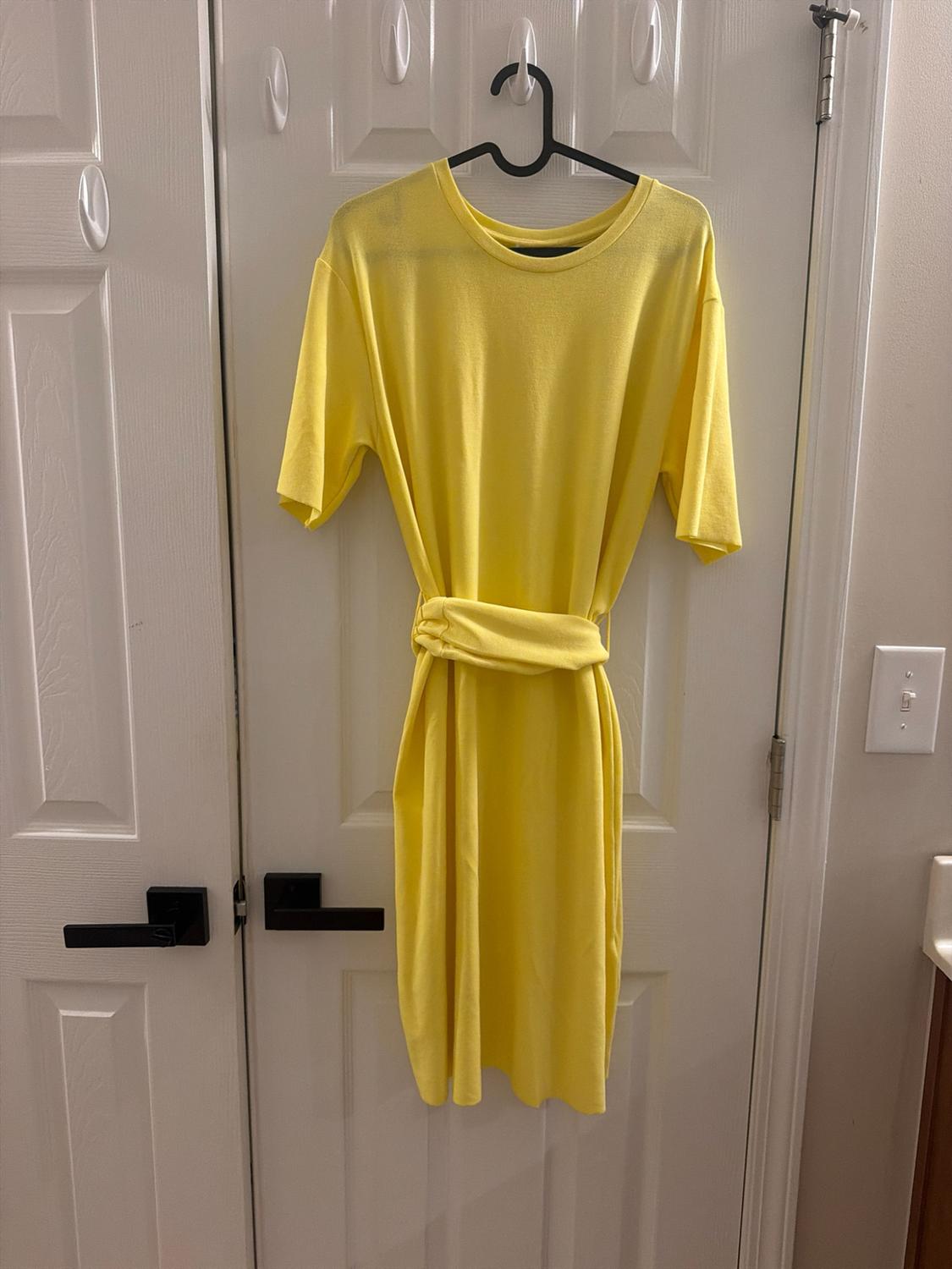 Short-sleeve yellow dress with a knotted waist detail, hanging on a black hanger on a white door.