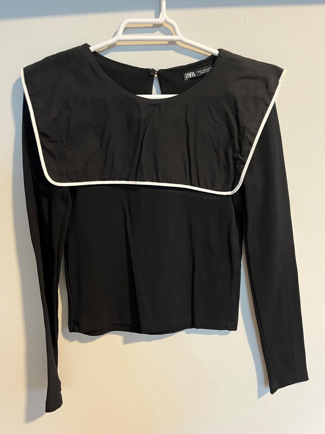 Black long-sleeved blouse with a square neckline and white trim, hanging on a white hanger against a light background.