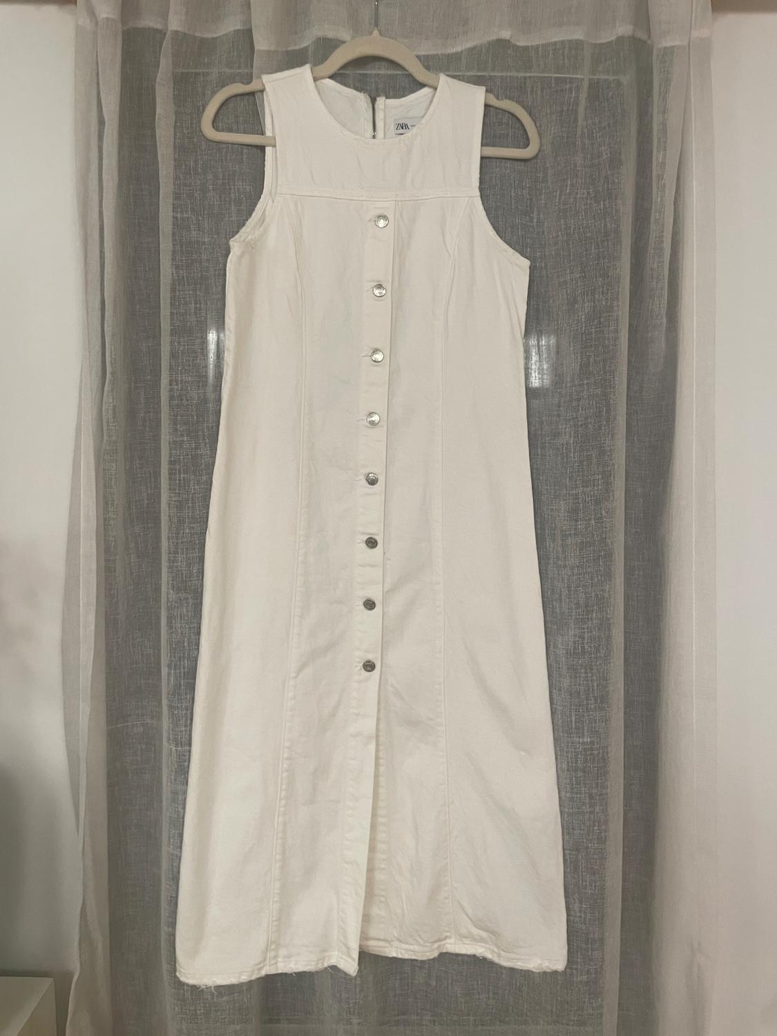 Sleeveless white dress with front buttons, hanging on a hanger in front of a semi-sheer curtain.
