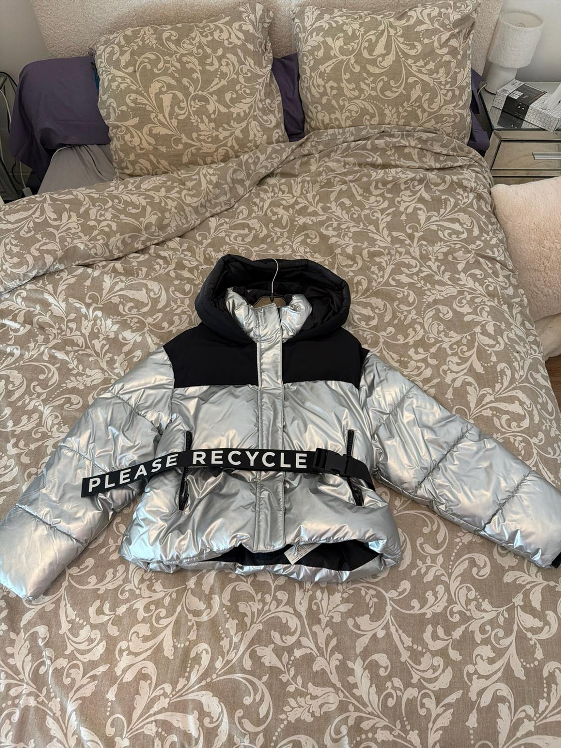Silver and black padded jacket on a bed with patterned sheets. It has a black belt with the text PLEASE RECYCLE.