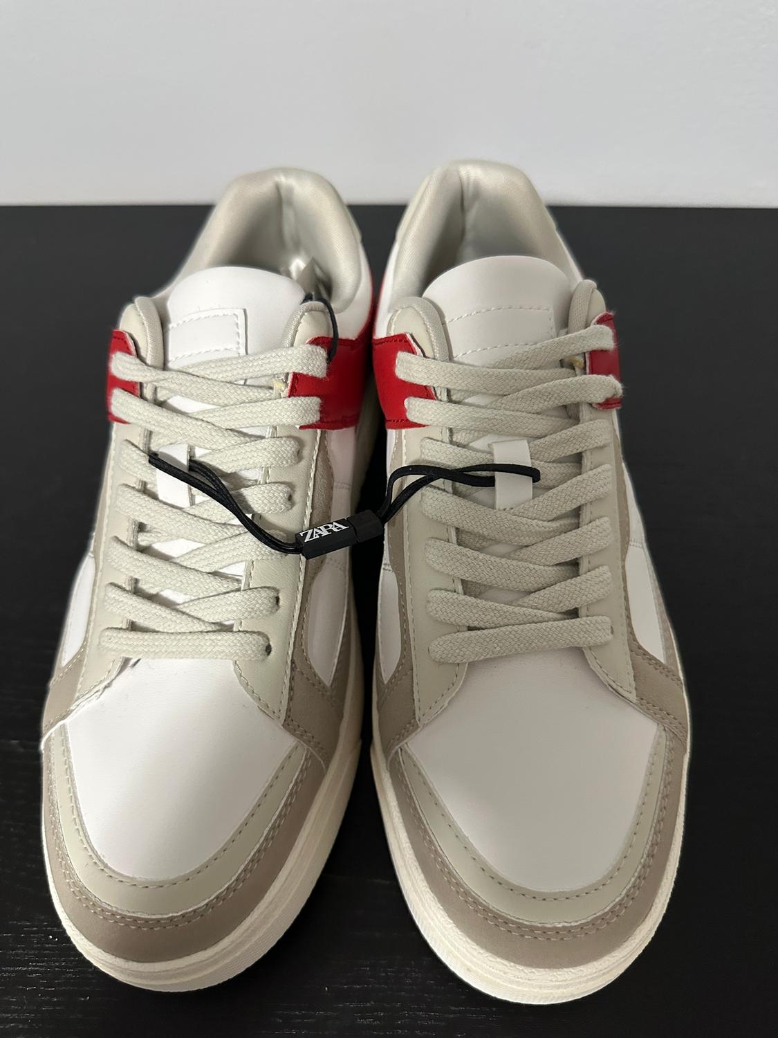 White and beige trainers with red detailing, beige laces, and a black label with "Zara" text, on a black background.