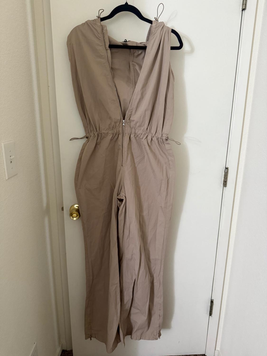 Beige jumpsuit without sleeves hanging on a black coat hanger over a white door. It has a front zip and waist adjustment.