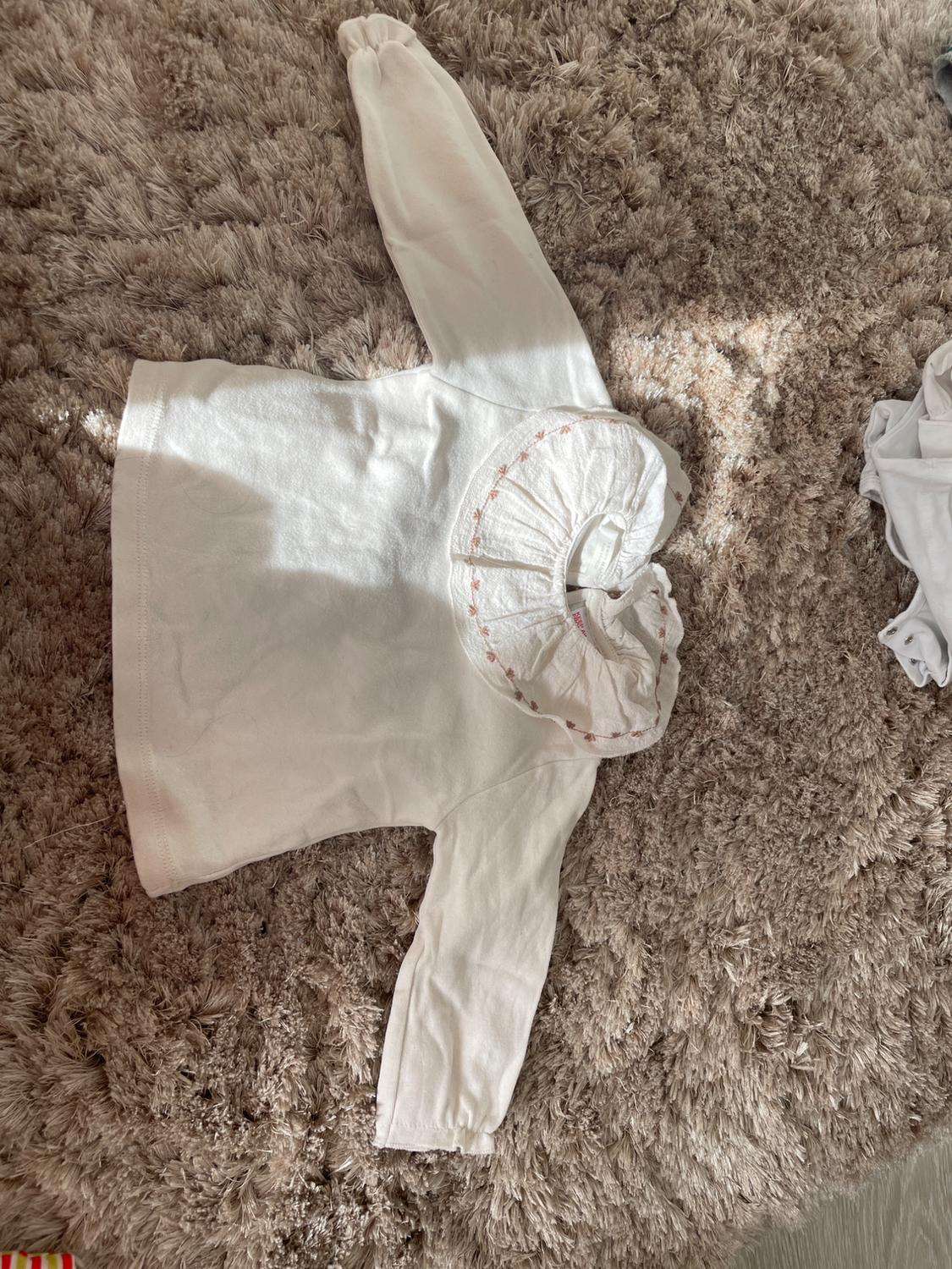 Long-sleeved white blouse with a ruffled collar and pink embroidery, placed on a beige, shaggy-textured rug.