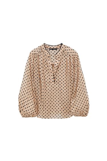 Beige blouse with black polka dots, puff sleeves and a V-neck. Black trim detail on the neck. White background.