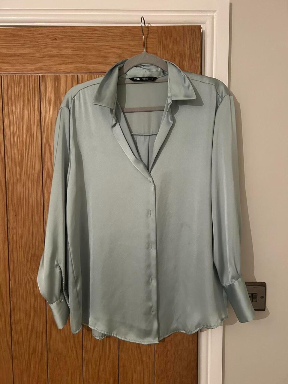 Light grey satin shirt hanging on a hanger over a wooden door. The shirt has a collar and front buttons.