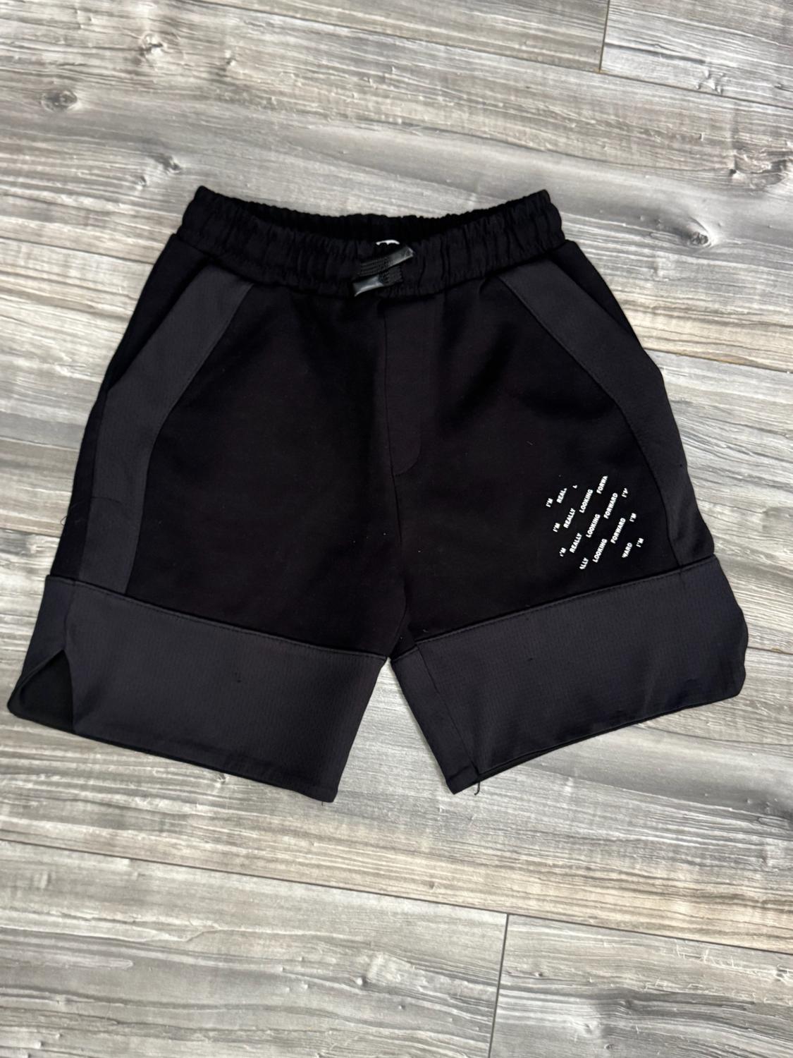 Black shorts on a grey wooden floor. They have an elasticated waistband, dark panels and the white text “I'M REALLY LOOKING FORWARD”.