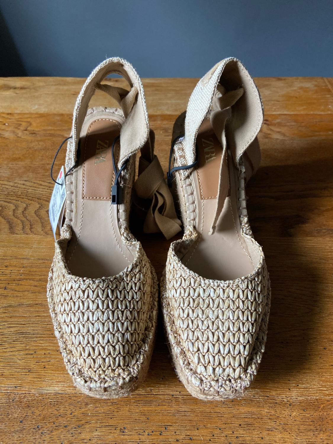 Pair of woven wedge espadrilles with ribbons on a wooden table. ZARA text visible on the insoles.