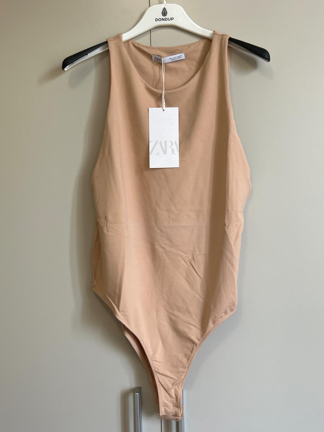 Beige sleeveless body hung on a white hanger with DONDUP text. Visible label with ZARA text. Wardrobe door background.