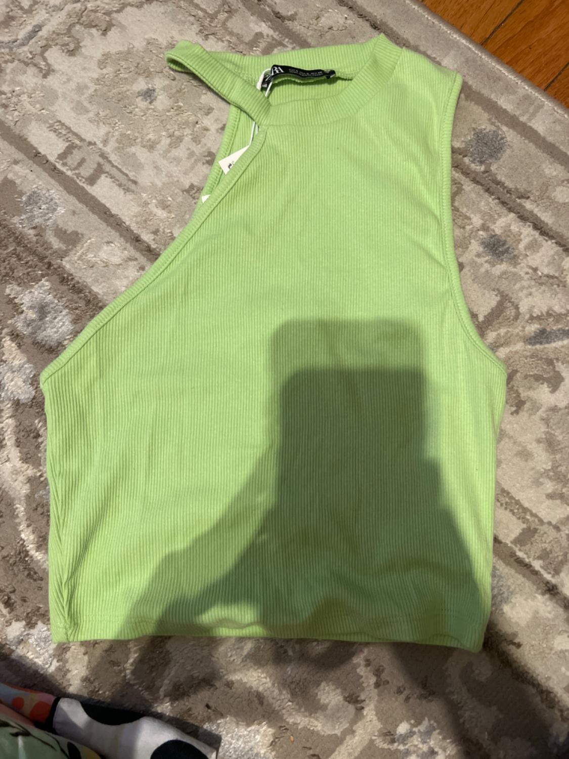 Sleeveless lime green ribbed knit top on a beige rug with geometric design, shadow of a phone visible in the center.