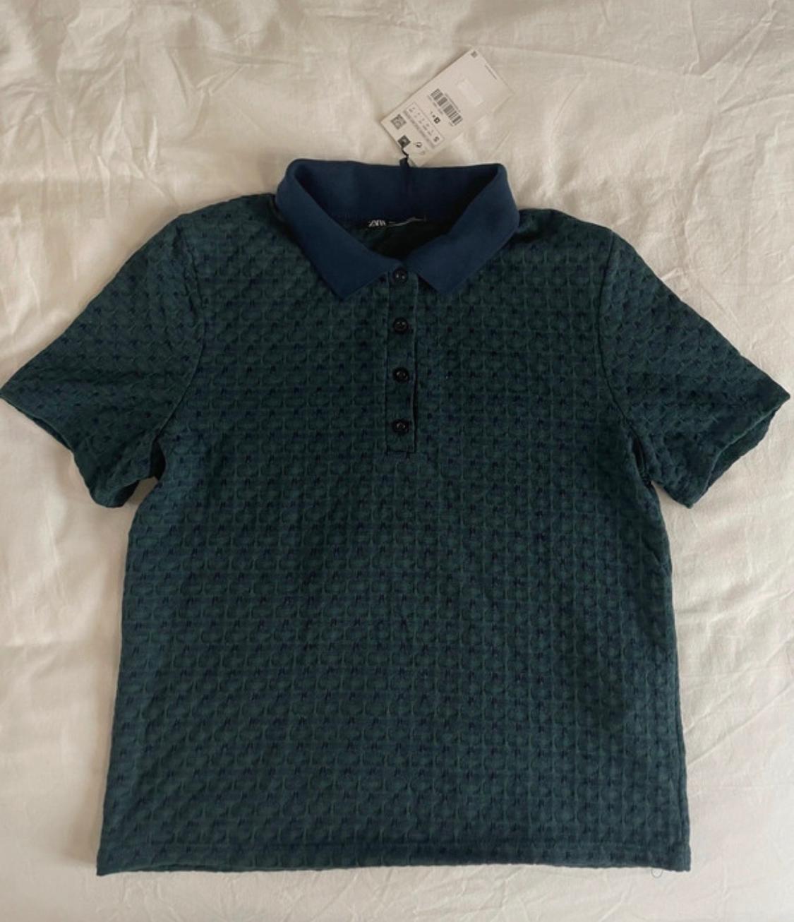Dark green short-sleeved polo shirt with textured fabric and a blue collar. The garment has a label and is on a crumpled white cloth.