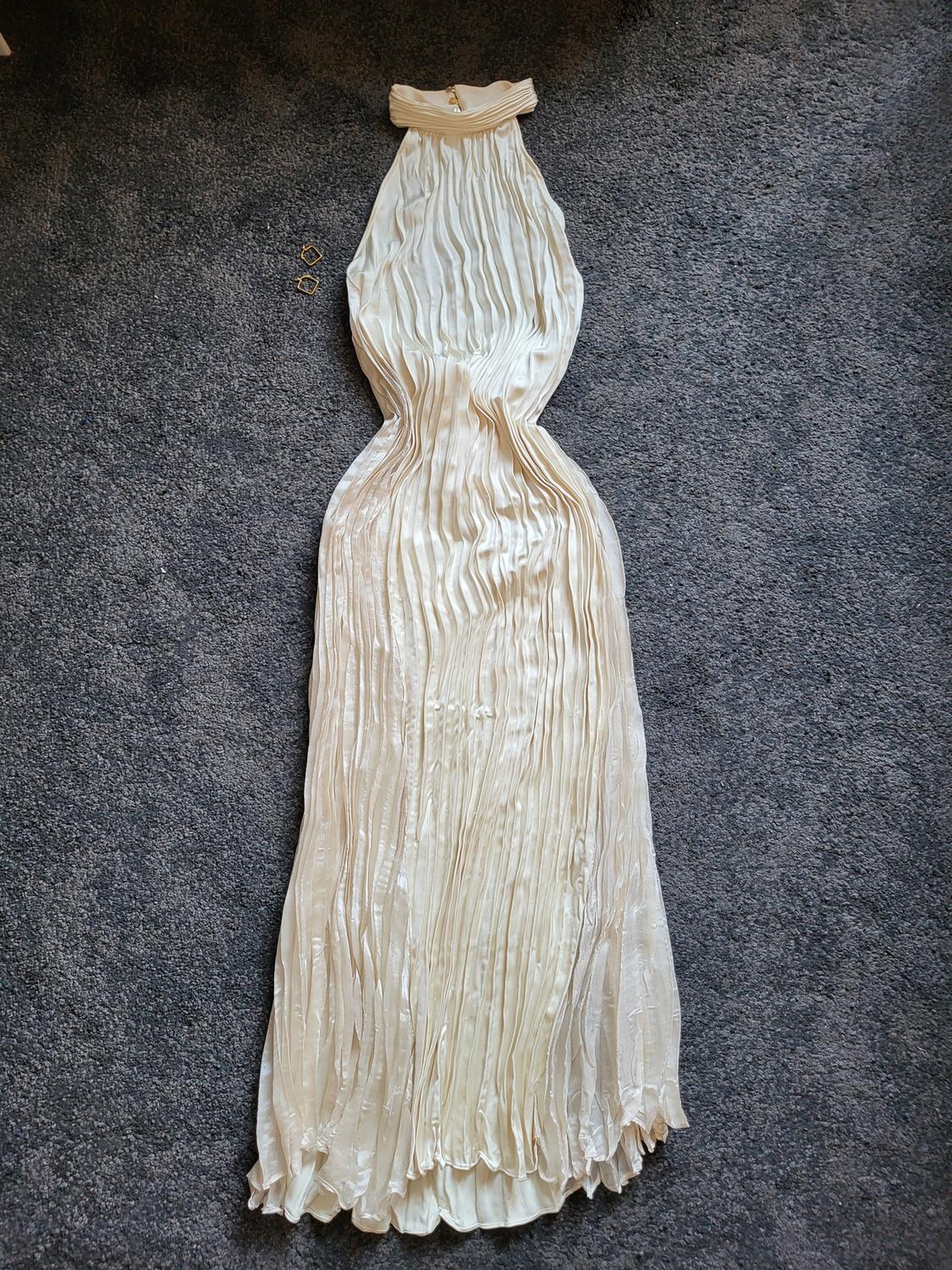 Pleated cream long dress on a grey rug, with a neck fastening. Two gold earrings on the left side.