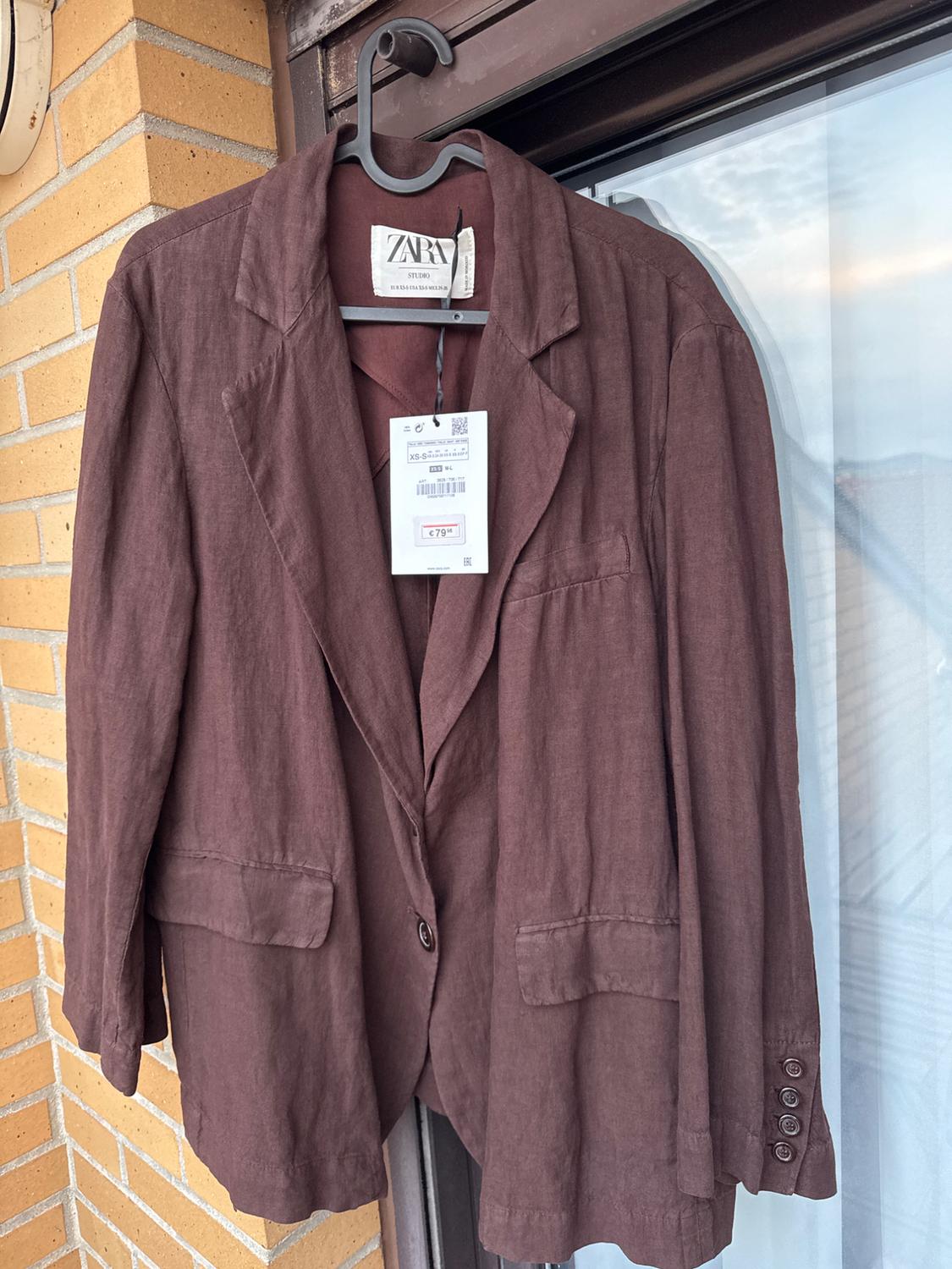 Brown jacket hanging on a coat hanger, with a visible price tag of £79.95. Brick wall and window in the background.