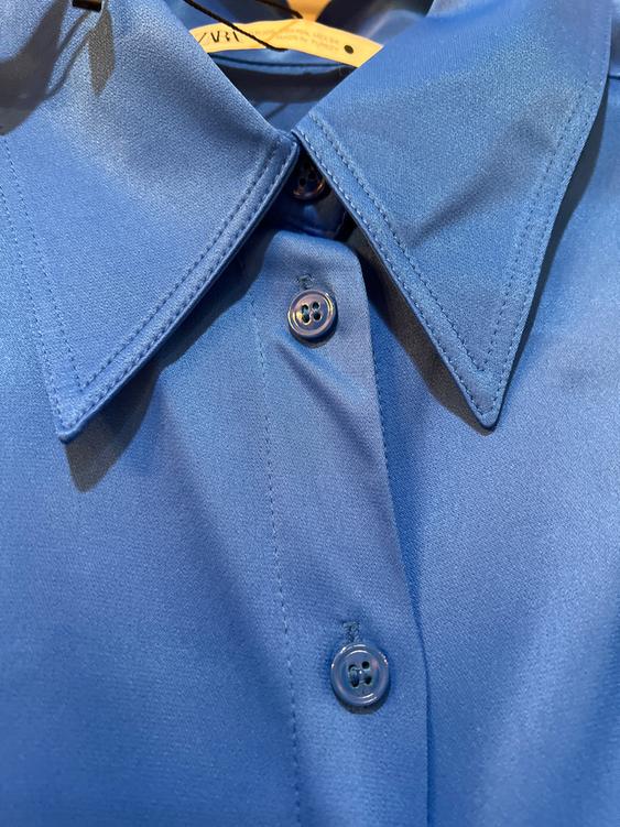 Blue satin fabric shirt with a wide collar and three visible buttons. Hanging tag at the bottom.