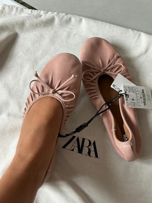 Pink ballet pumps with a bow, one worn on a foot and the other with the tag, on a white fabric with the text ZARA.