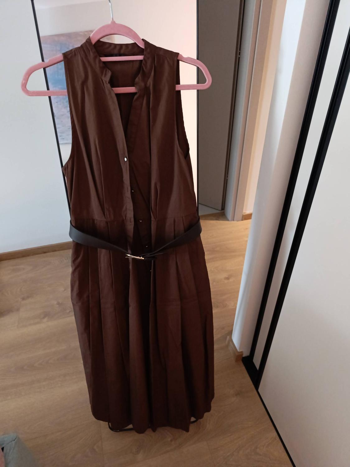 Brown sleeveless shirt dress with a dark belt and pleated skirt, hanging on a pink hanger in a room with a wooden floor.