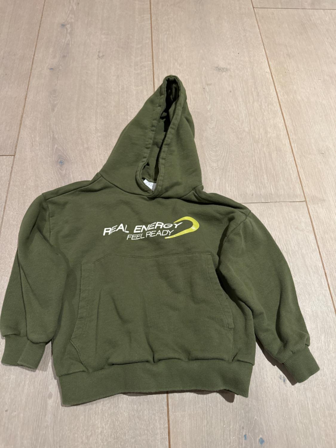 Green hooded sweatshirt on a wooden floor. Text on the front: "REAL ENERGY FEEL READY". Large front pocket.