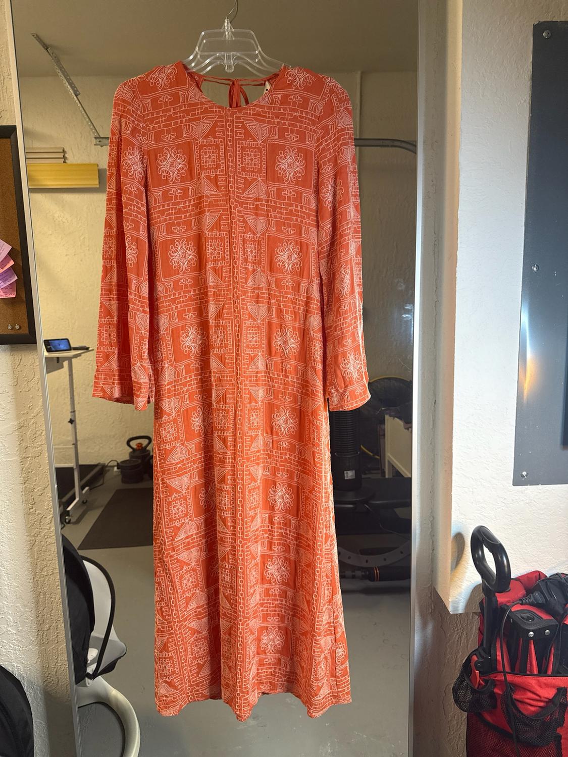 Long orange dress with a white geometric print hanging on a hanger in front of a mirror reflecting a room.