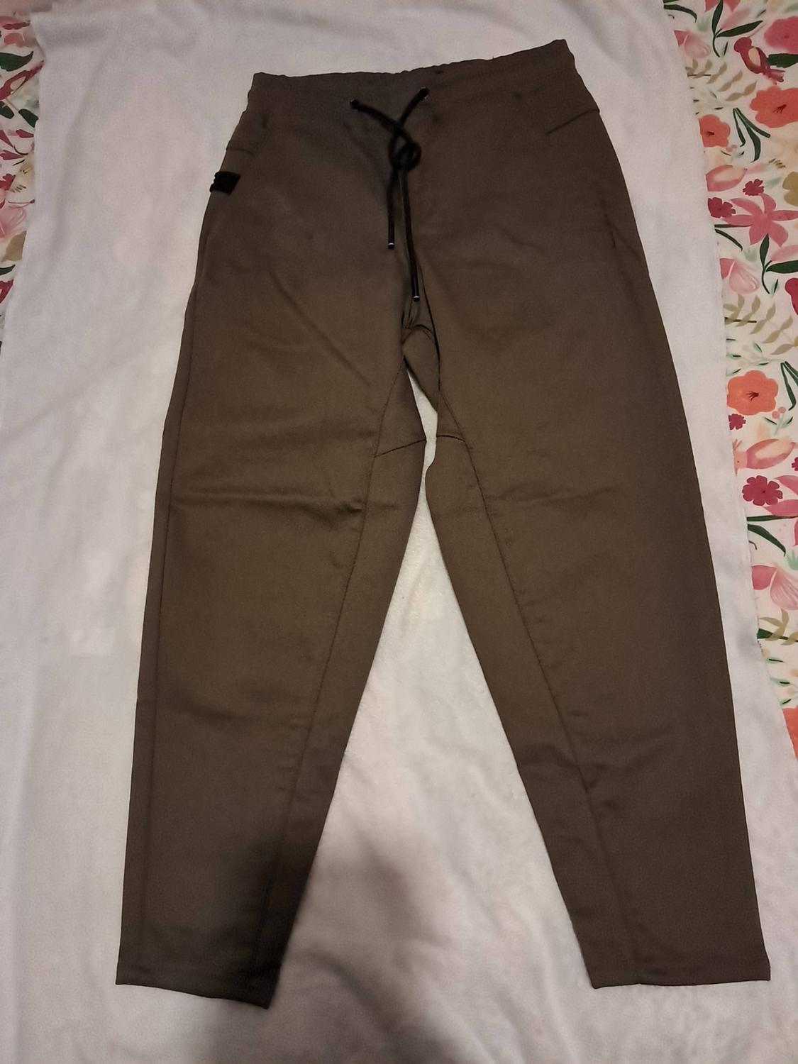 Brown trousers with an elasticated waist and black drawstring, laid on white fabric with a floral-patterned background.