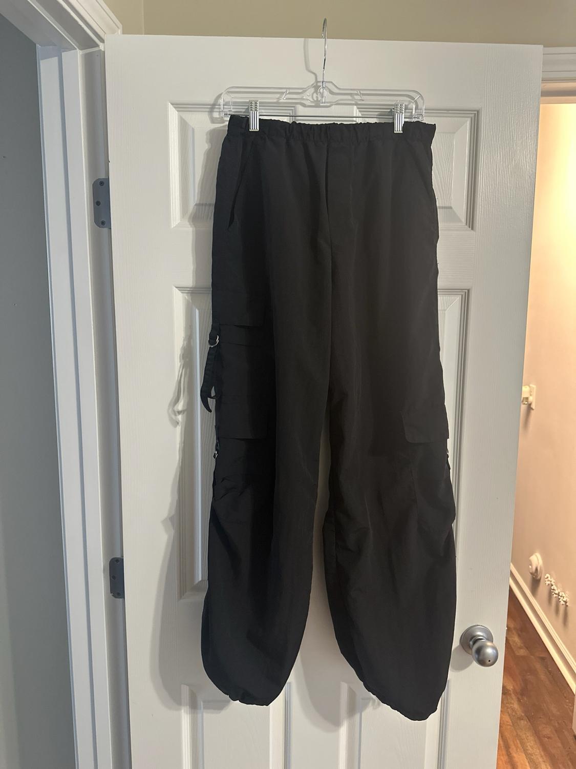Wide black cargo trousers with side pockets and an elasticated waist, hanging from a transparent hanger on a white door.