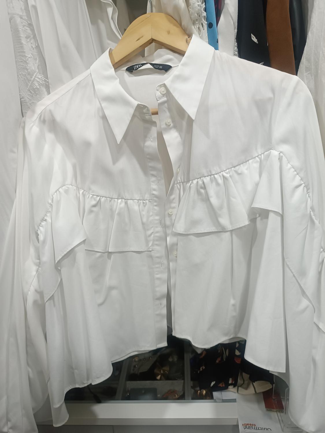 White blouse with ruffled sleeves and a classic collar, hanging on a wooden hanger in a wardrobe full of assorted clothes.