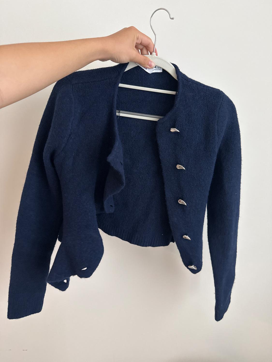 A hand holds a navy blue knit cardigan with silver buttons on a white hanger against a light-colored wall. Casual style.