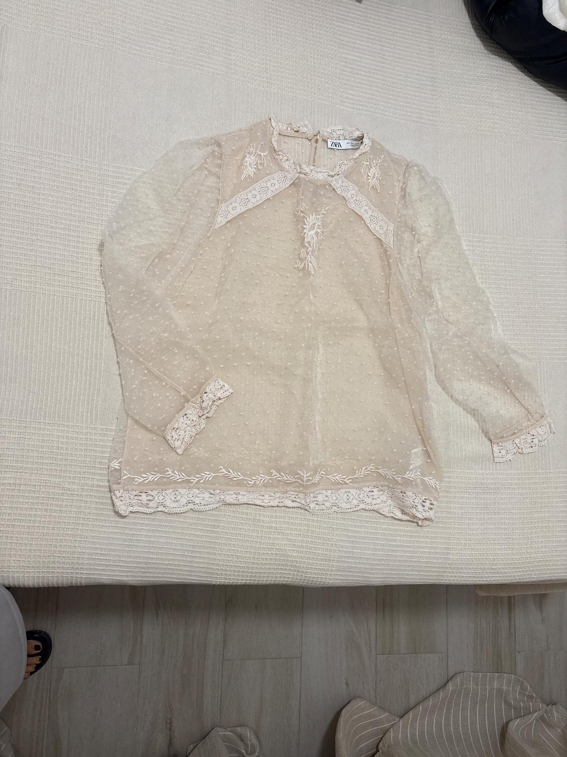 Beige lace blouse with floral details and sheer long sleeves, placed on a light surface. Visible label.