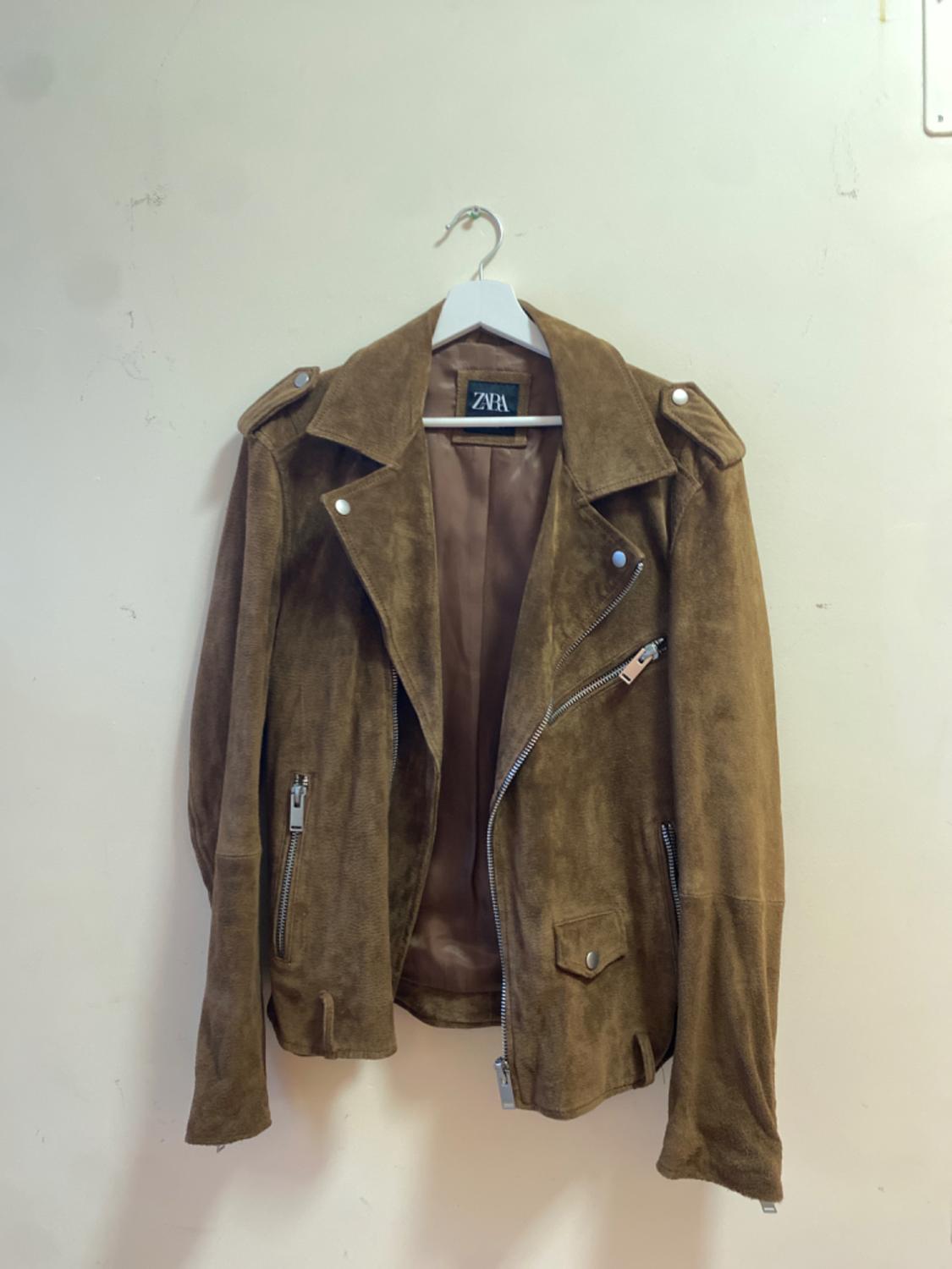 Brown suede jacket with an asymmetrical zip and lapels, hanging on a white hanger. Visible label: ZARA. White background.