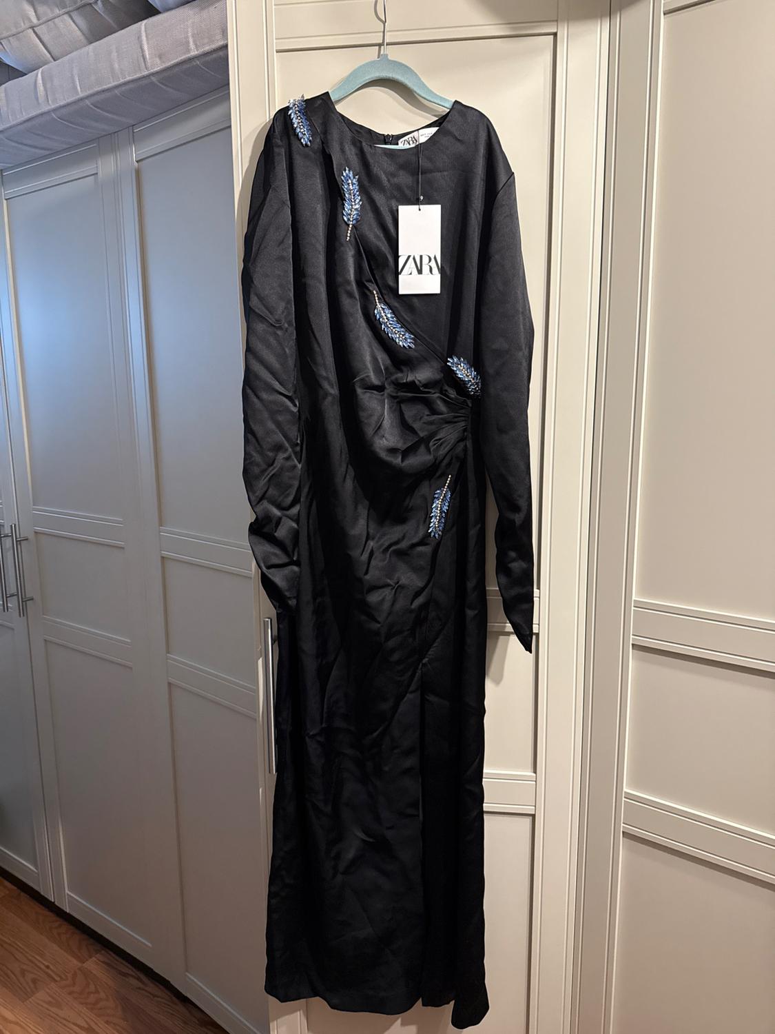 Black dress with blue embroidery hanging on a blue hanger. Visible tag with the text "ZARA". White wardrobe background.