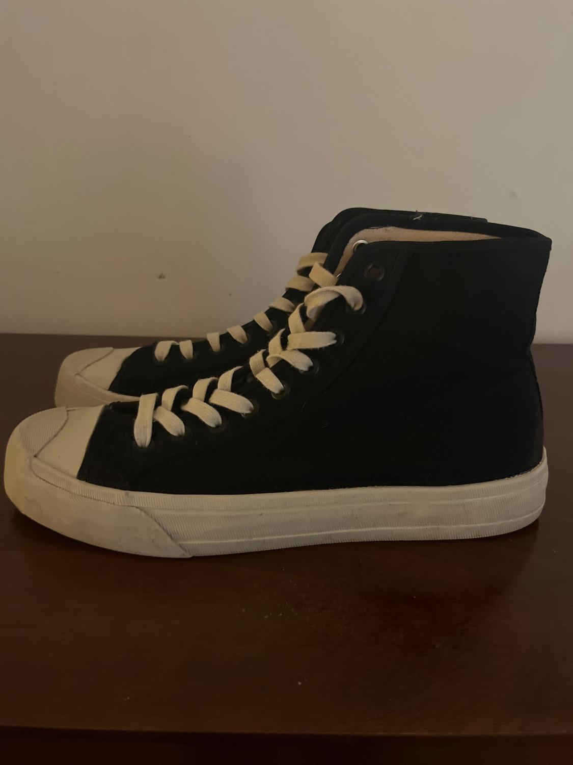 Black canvas trainers with white laces on a wooden surface, white background. Casual and classic style.