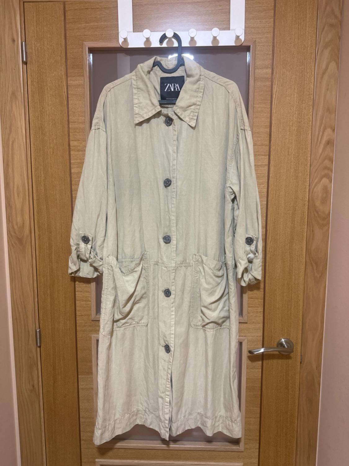 Beige coat hanging on a wooden door. It has large buttons, front pockets, and a visible label with the text "ZARA".