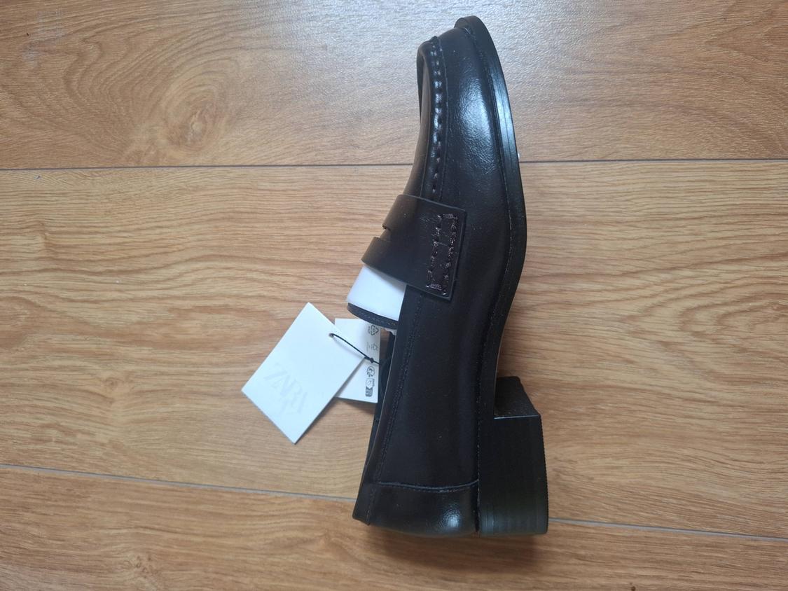 Black loafer shoe resting on a wooden floor. It has a white label with the text ZARA.