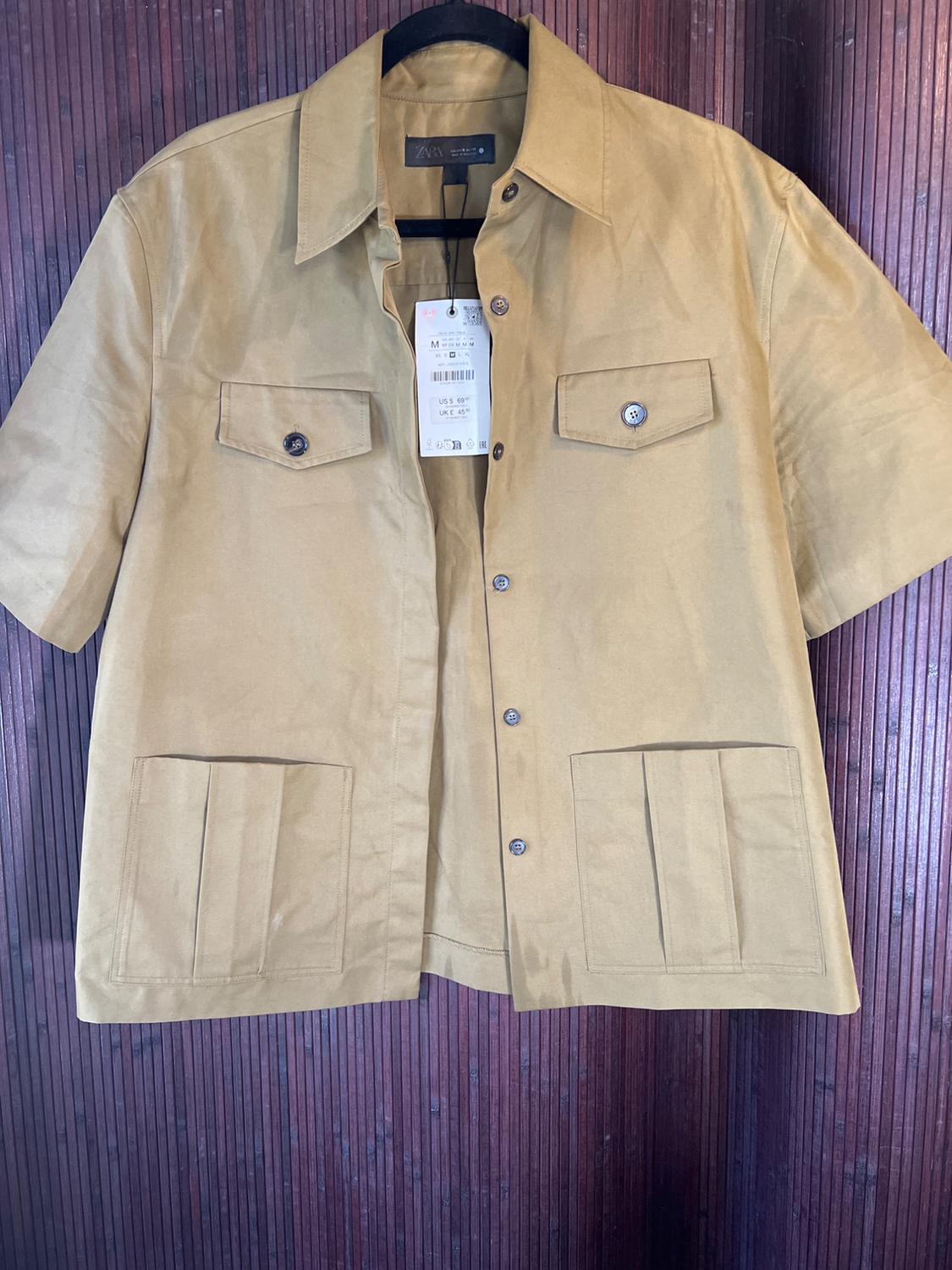 Beige short-sleeved shirt with front pockets and hangtag, hanging on a dark wooden wall.