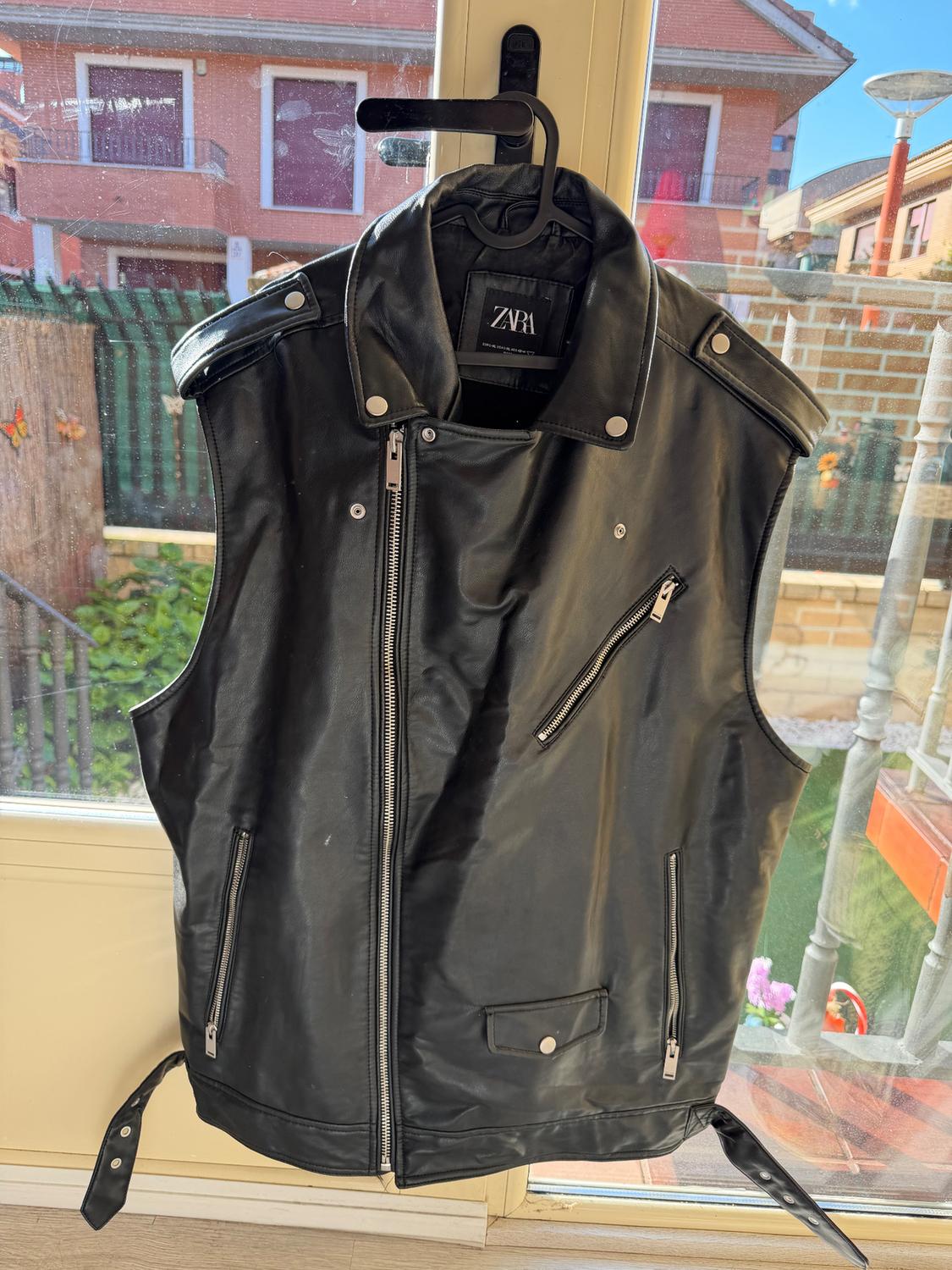 Black leather waistcoat hanging on a hanger in front of a window. Visible label: "ZARA". Background with houses and blue sky.