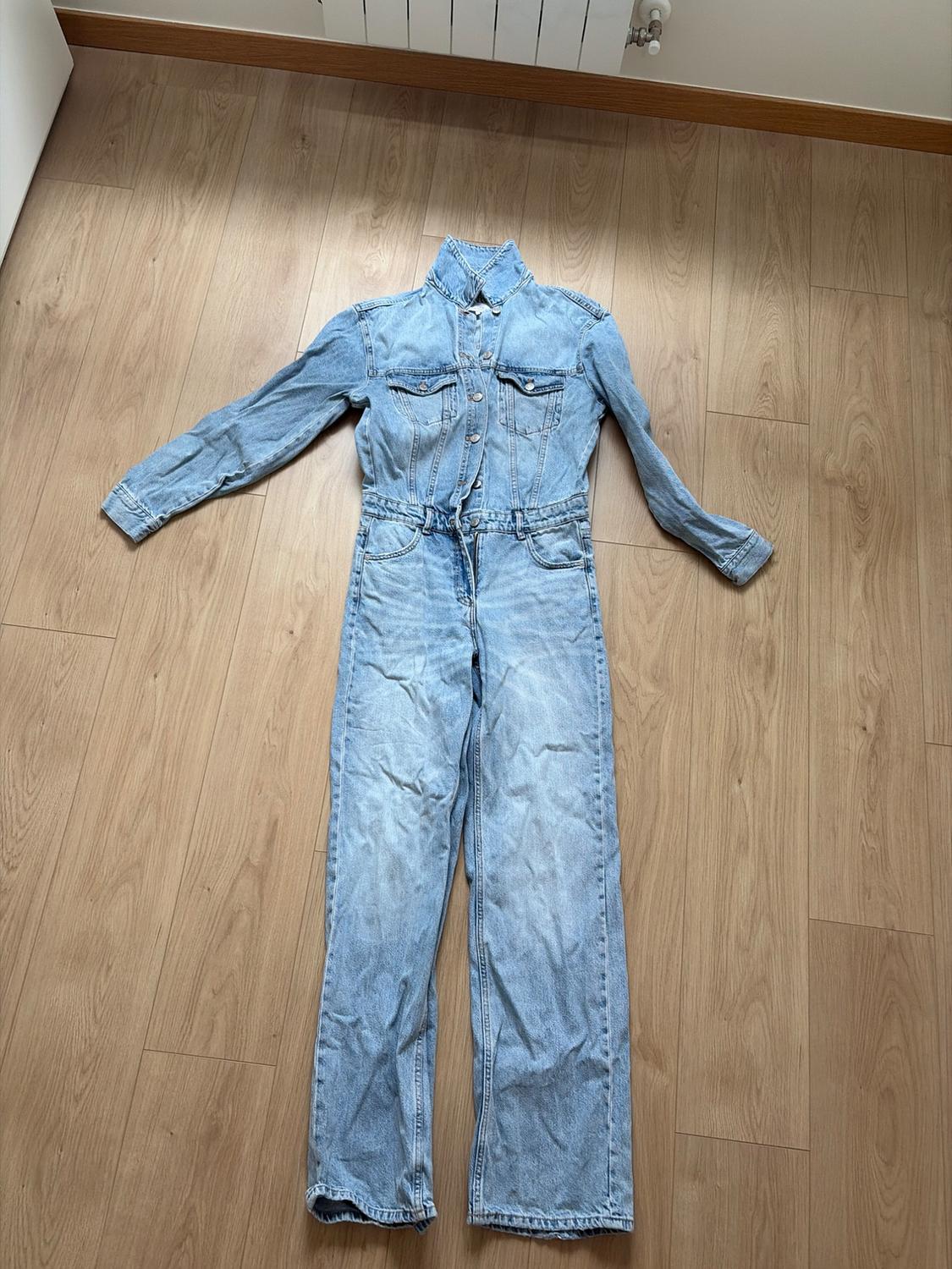 Light blue, long-sleeved denim jumpsuit with front buttons, laid out on a light wooden floor. Casual and relaxed style.