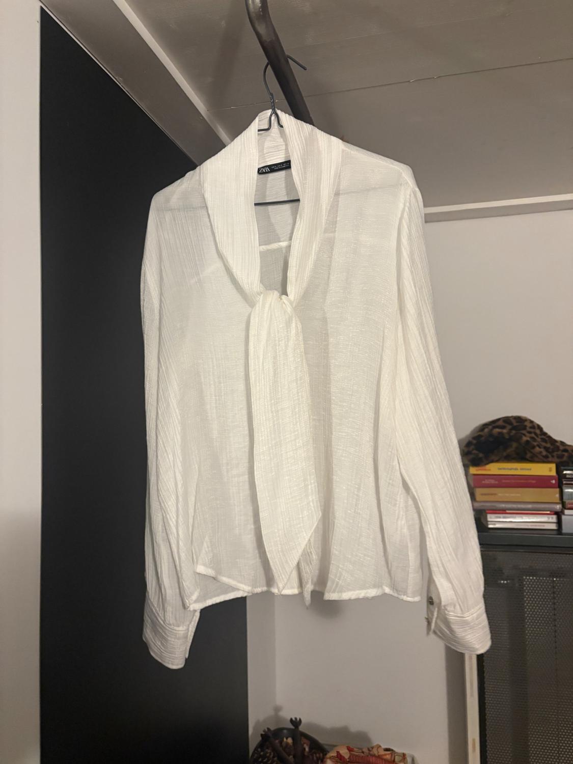 White blouse made of light fabric, hanging on a hanger in a wardrobe. Background with stacked books and neutral-toned decor.