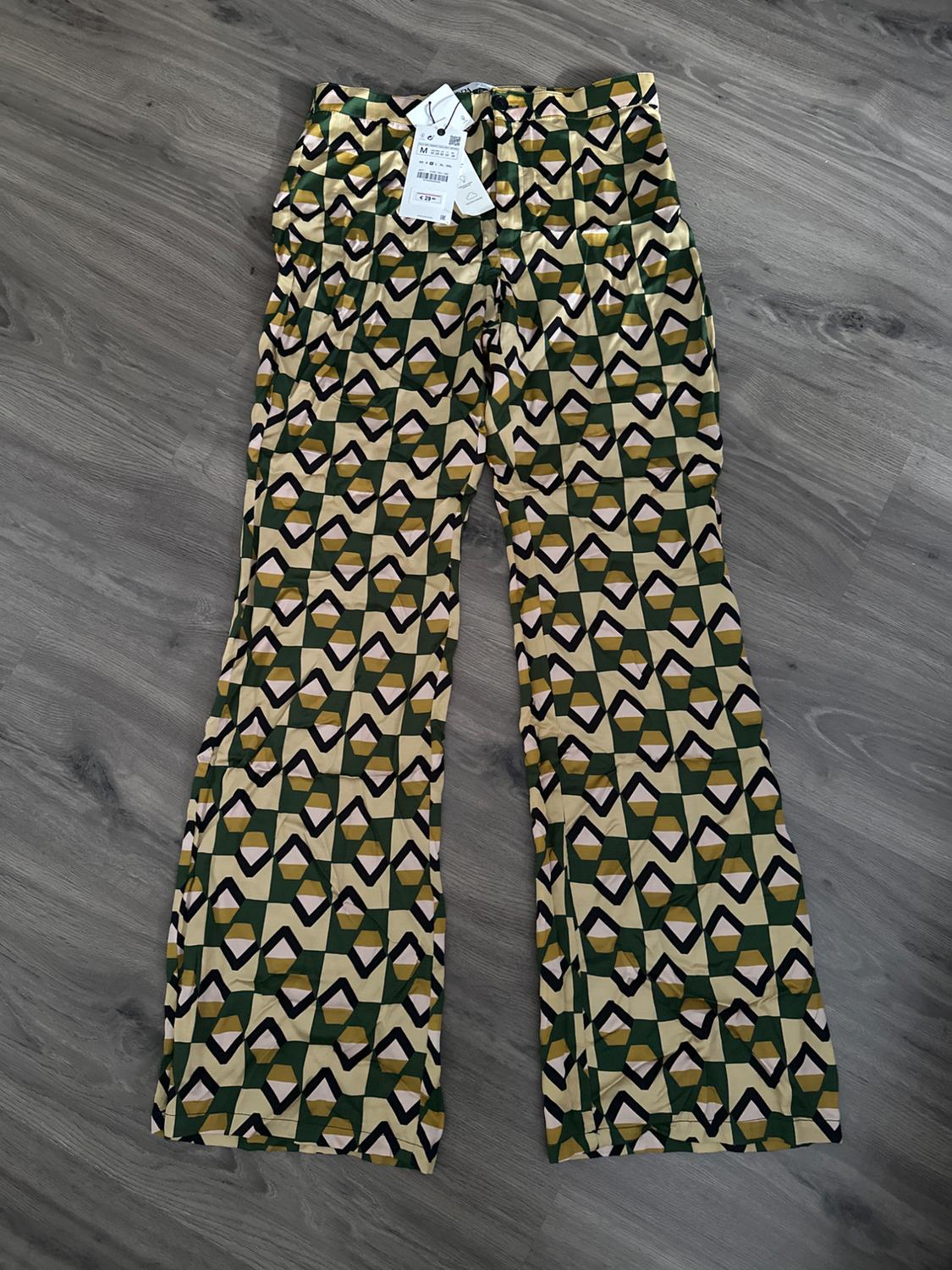Geometric patterned trousers in shades of green and beige on a grey wooden floor. Tags hanging from the waist.
