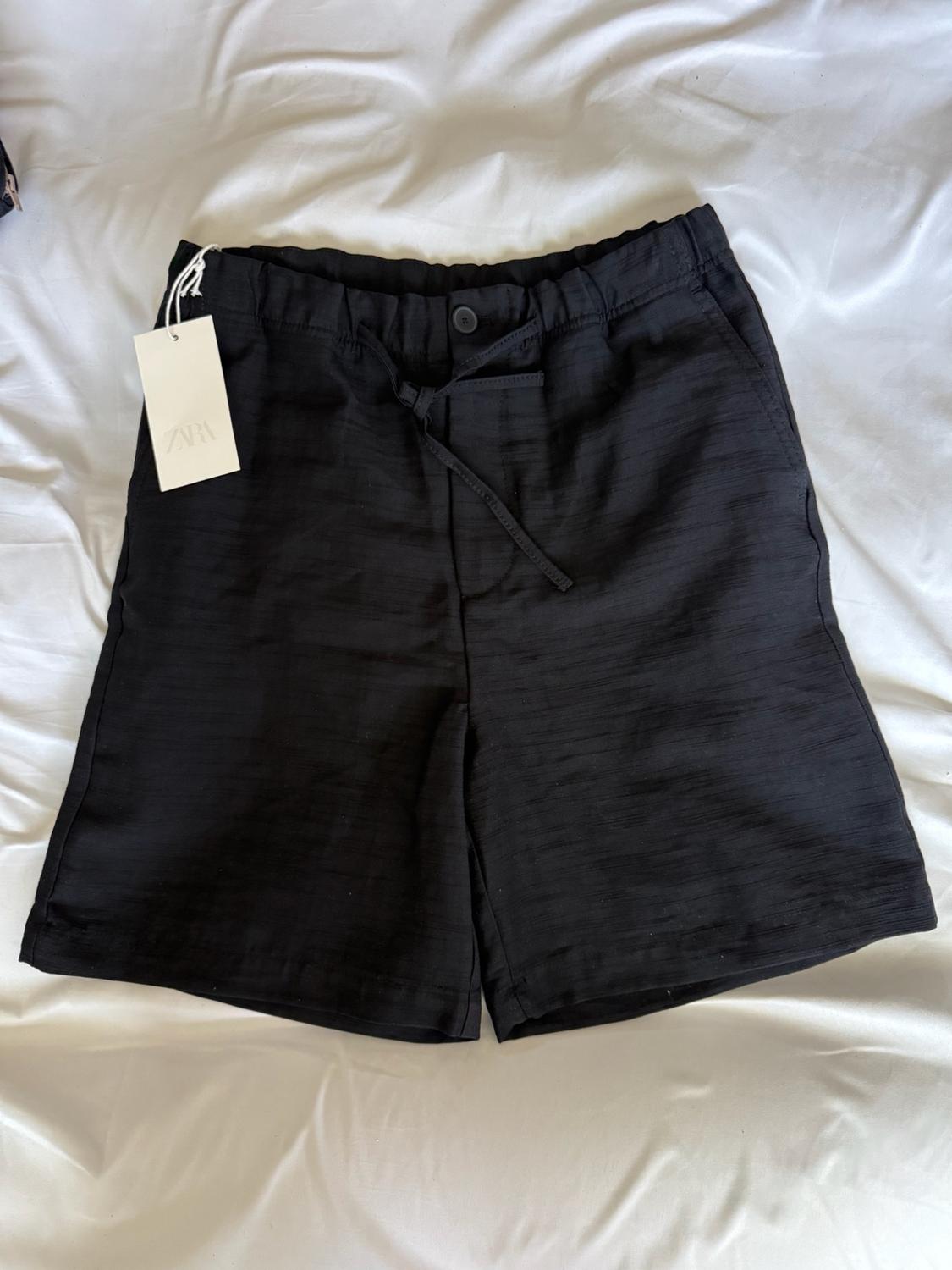 Black shorts with an elasticated waistband and drawstring, a label bearing the text ZARA, on a crumpled white sheet.