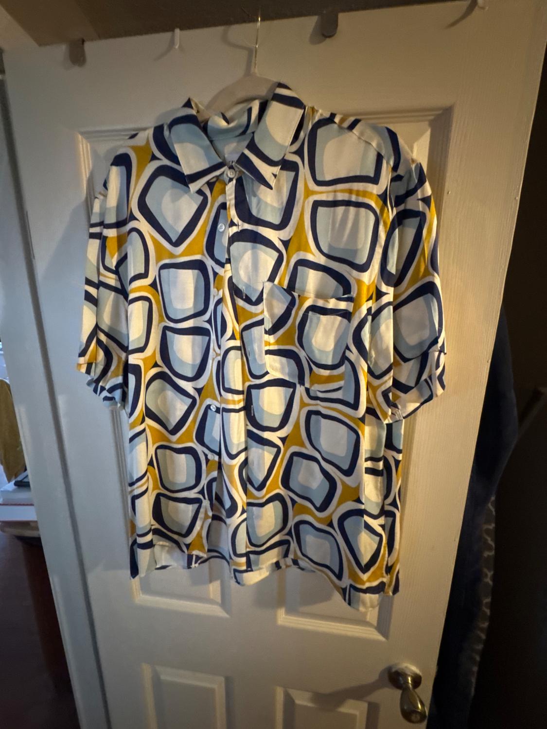 Short-sleeved shirt with pocket and geometric print in blue, white, and yellow, hanging on a white door.